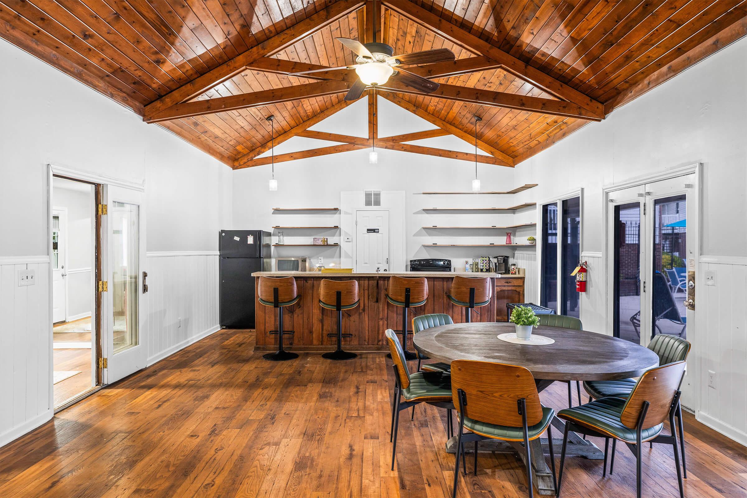 A spacious kitchen and dining area featuring a high wooden ceiling with exposed beams, modern bar stools at the counter, a circular dining table with wooden chairs, and various kitchen appliances. The floor is made of polished wood, and there are shelves against the walls for storage.