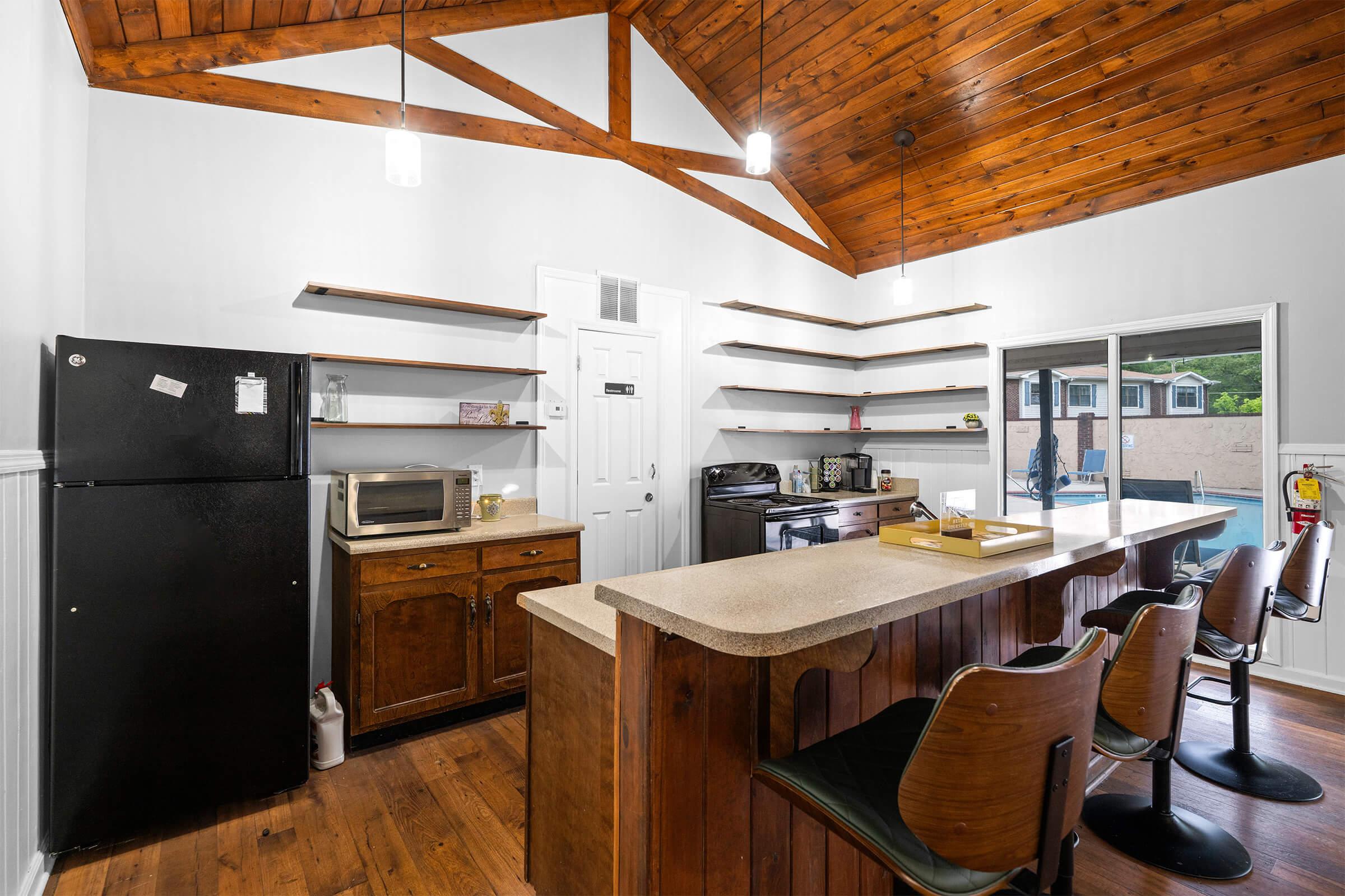 A modern kitchen featuring dark wood shelves, a black refrigerator, a microwave, and a gas stove. The kitchen has wooden flooring and a high ceiling with wooden beams, illuminated by pendant lights. There is a countertop with four bar stools and a sliding glass door leading to an outdoor pool area.
