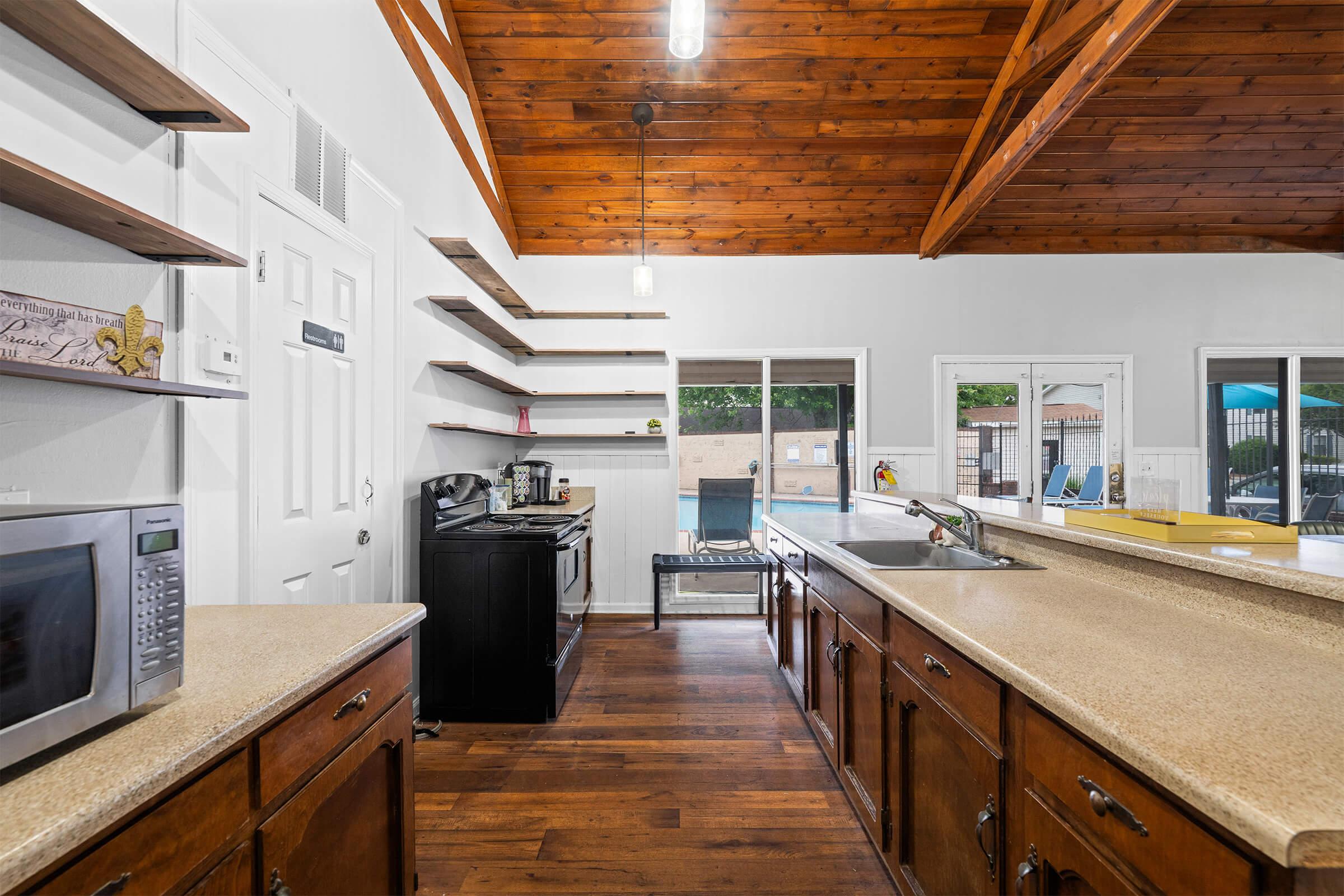 Modern kitchen with wooden beams and shelves, featuring a countertop, microwave, and a black stove. Large windows provide a view of an outdoor pool area. The floor is dark wood, creating a warm atmosphere. Sunlight illuminates the space, suggesting a bright and inviting environment.