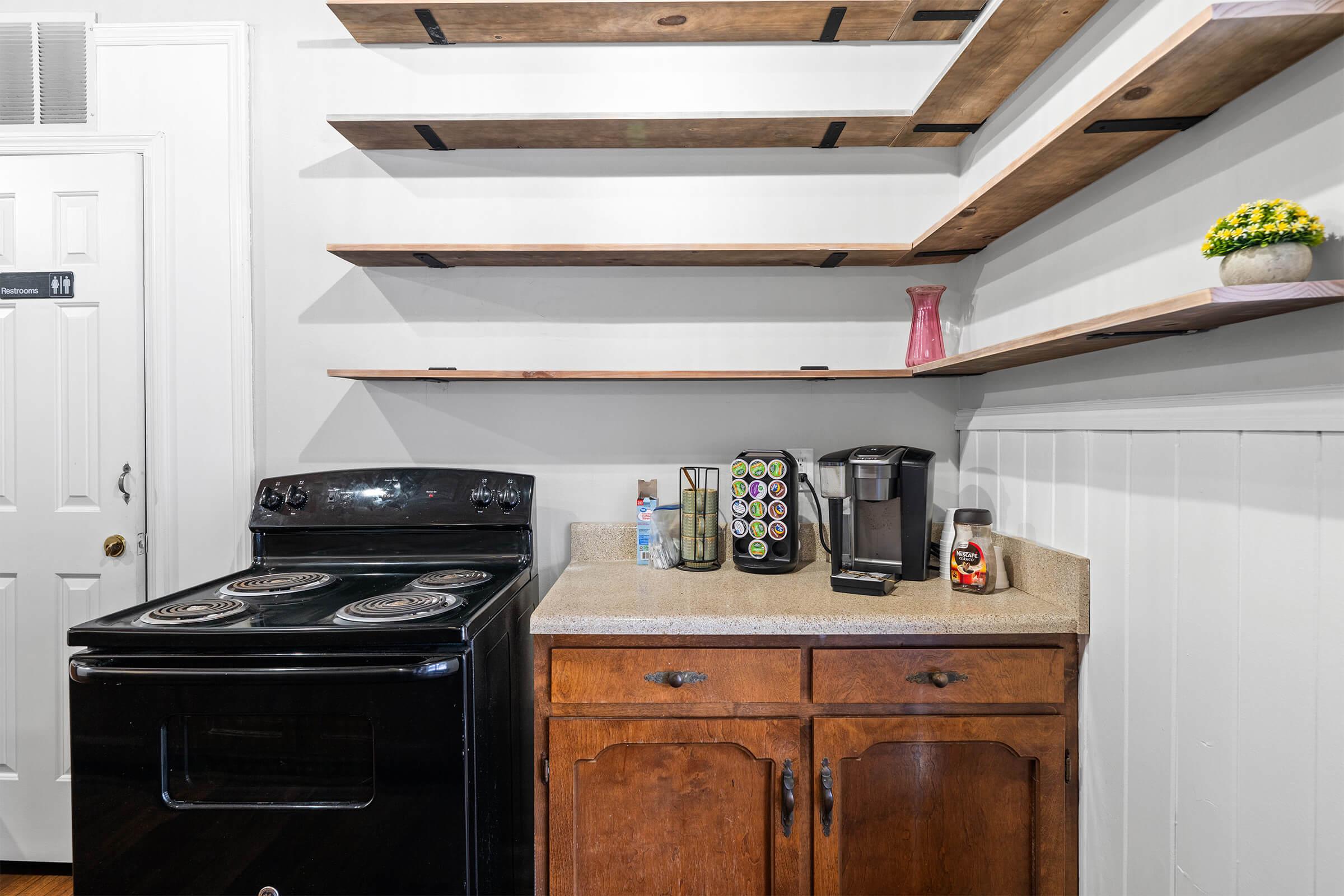 A modern kitchen with a black stove and dark wooden cabinets. Above the counter, there are several wooden shelves displaying decorative items. A coffee maker and a green and brown canister sit on the countertop. Natural light highlights the space, creating a welcoming atmosphere.