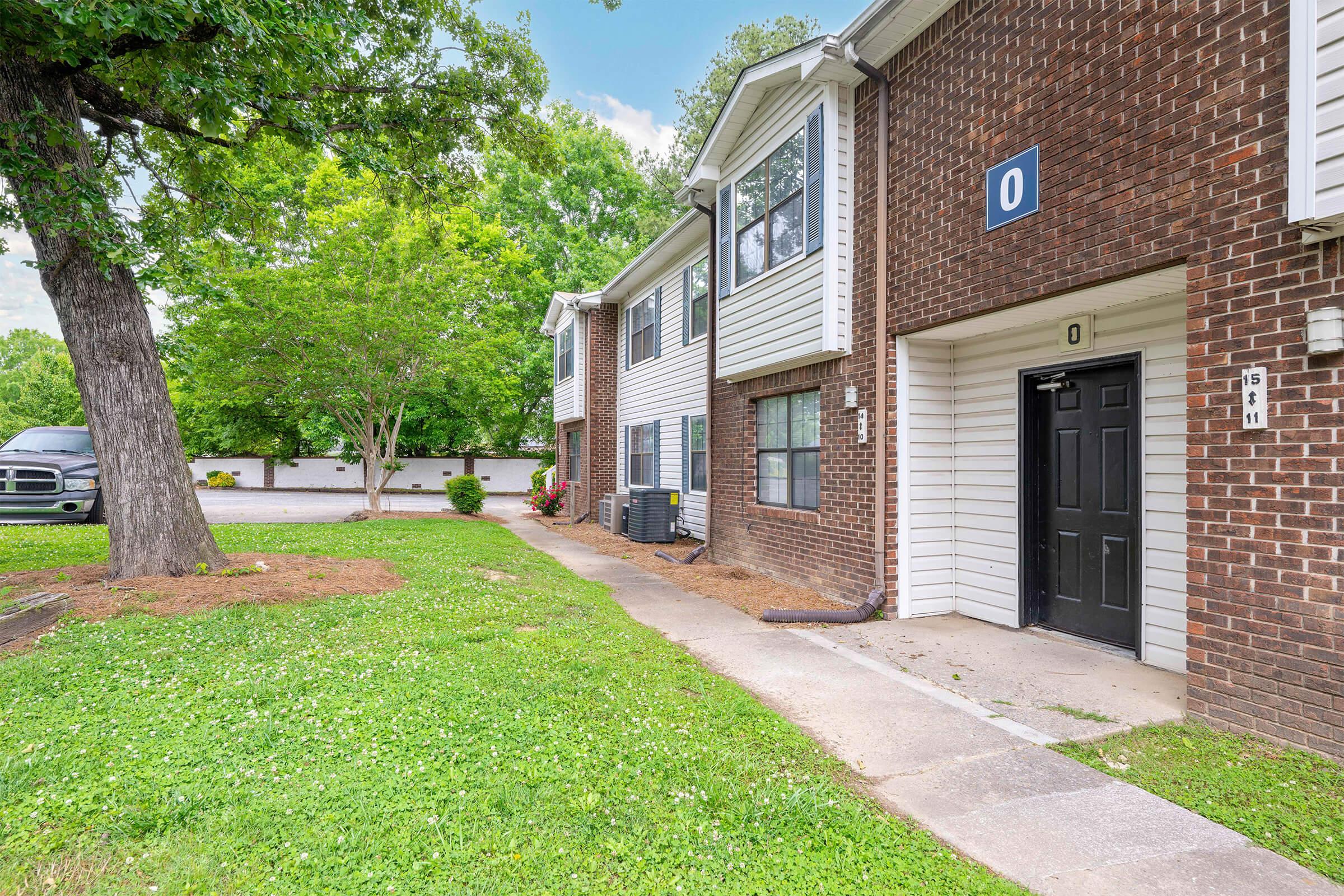 A brick apartment building with numbered units, featuring a pathway leading to a black door. Surrounding greenery includes trees and well-maintained lawn areas. A parking lot is visible in the background, with a truck parked nearby. The scene is bright and inviting, suggesting a residential neighborhood.
