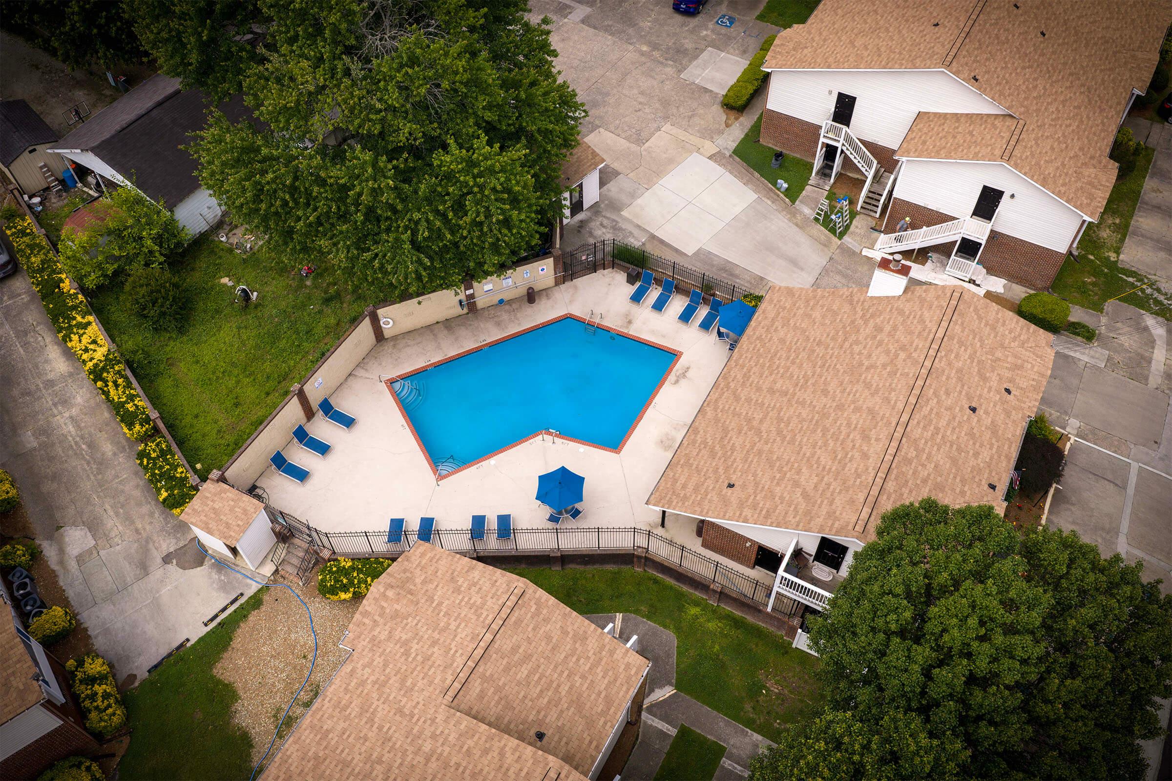 Aerial view of a residential area featuring a swimming pool with blue lounge chairs and an umbrella. The pool is surrounded by a fence, with several single-story houses visible nearby, surrounded by greenery and paved pathways.