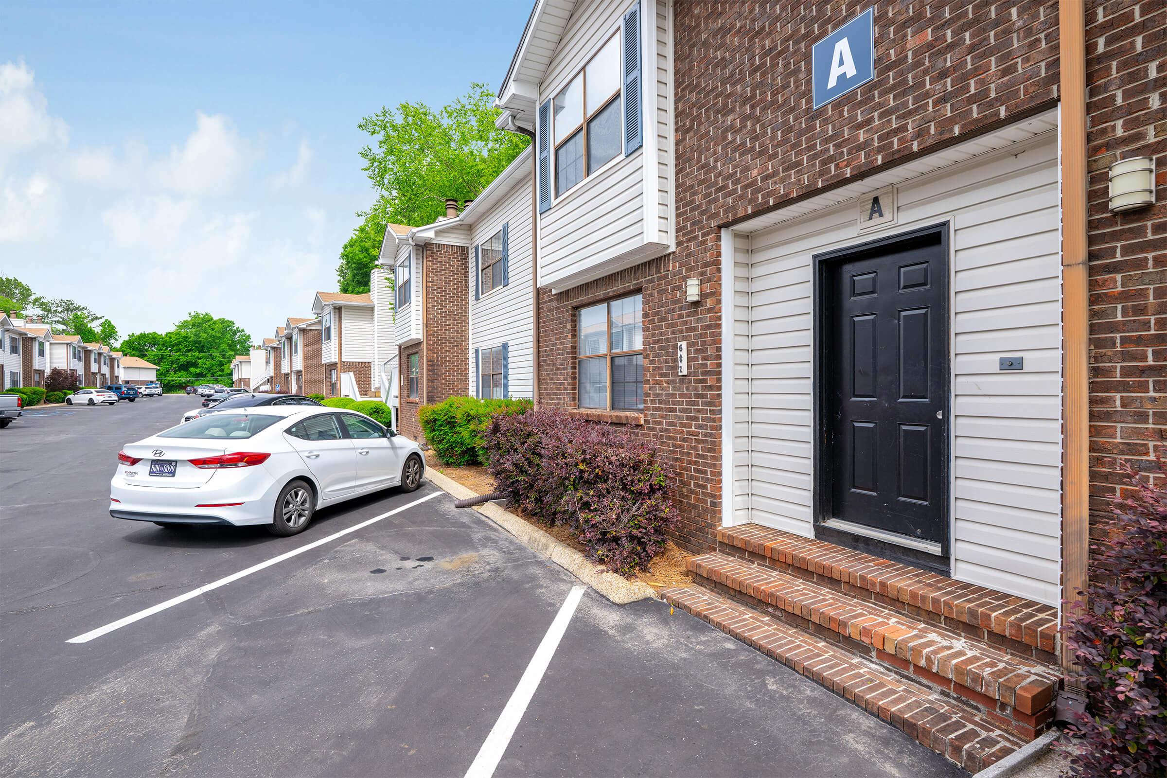 Exterior view of a residential building with a dark front door labeled "A." A white car is parked in front of the building, which features brick and siding. Lush green trees and shrubs are visible in the background, indicating a pleasant environment. The scene captures a well-maintained apartment complex.