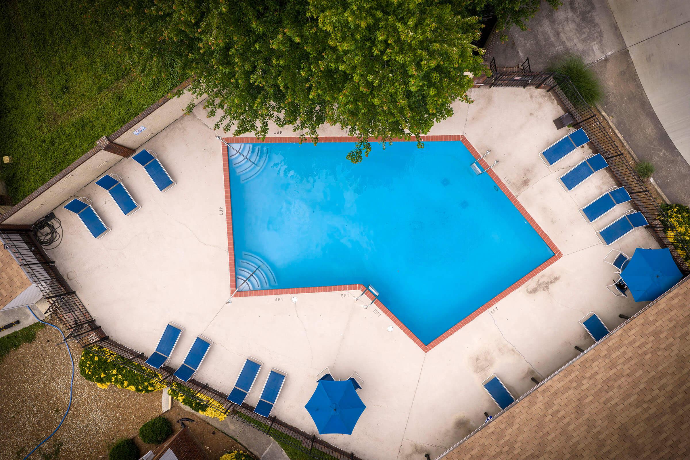 Aerial view of a rectangular swimming pool surrounded by lounge chairs and umbrellas. The pool features a clear blue water surface, and there are grassy areas on one side, with a paved pathway and a few trees nearby. The scene is bright and inviting, ideal for relaxation and leisure.