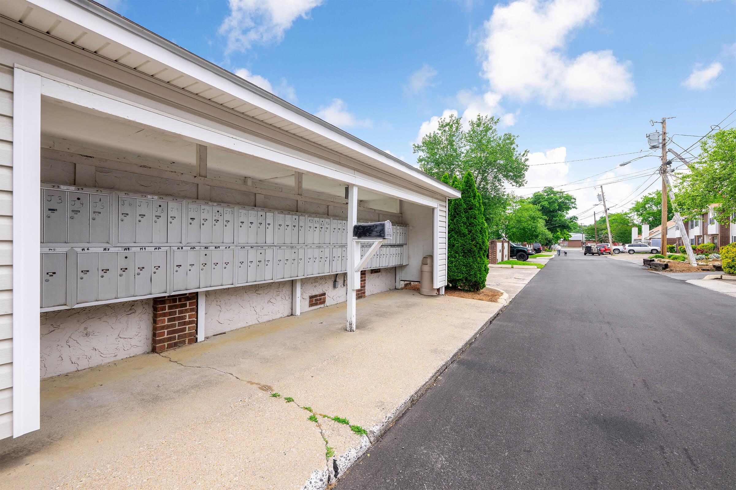 A row of individual mailboxes under a covered structure, located beside a paved walkway. Trees and landscaping are visible in the background along a quiet street lined with power poles and neighboring houses. The sky is partly cloudy, creating a bright and inviting atmosphere.