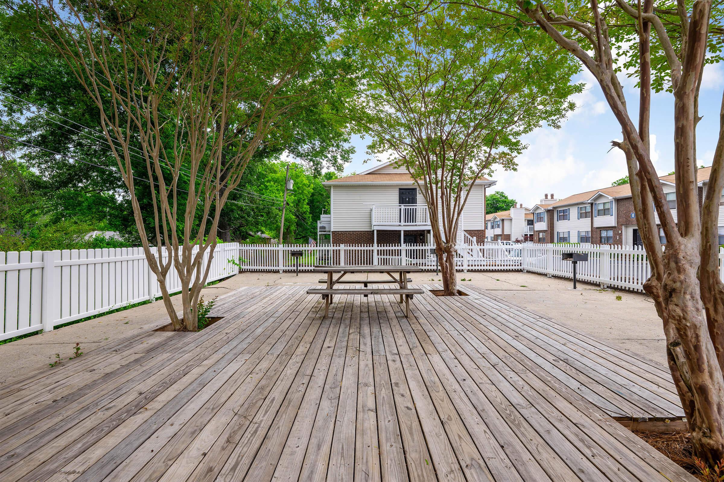 A wooden deck area with a picnic table situated under leafy trees. The space is enclosed by a white fence, and residential buildings are visible in the background. The setting is bright and inviting, featuring a clear sky and greenery.