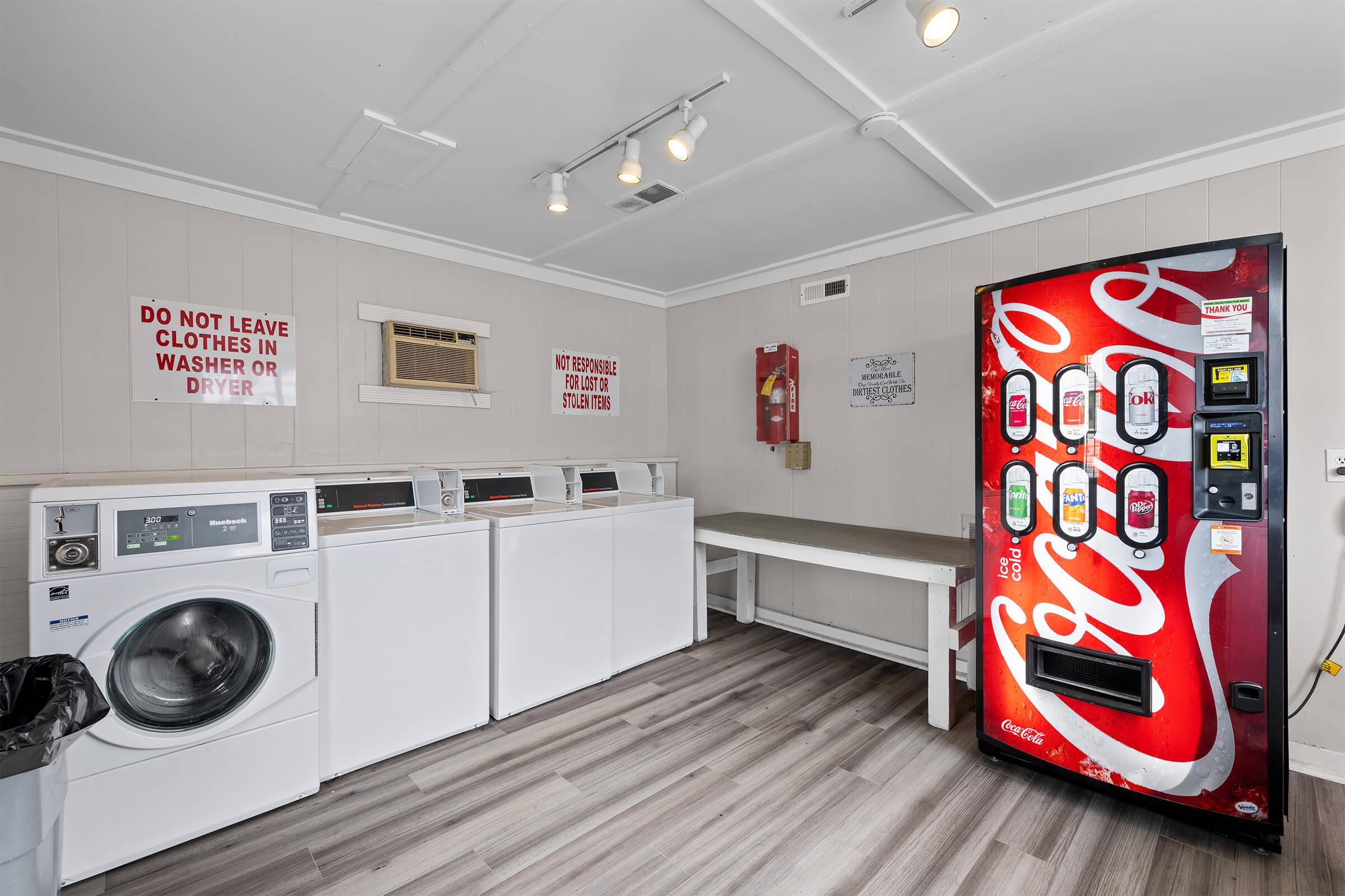 Laundry room featuring three front-loading washing machines and a dryer. A Coca-Cola vending machine is present on the right. The walls are adorned with signs instructing patrons not to leave clothes in the washer or dryer, and other operational notices. The room has a clean, modern aesthetic with laminate flooring.