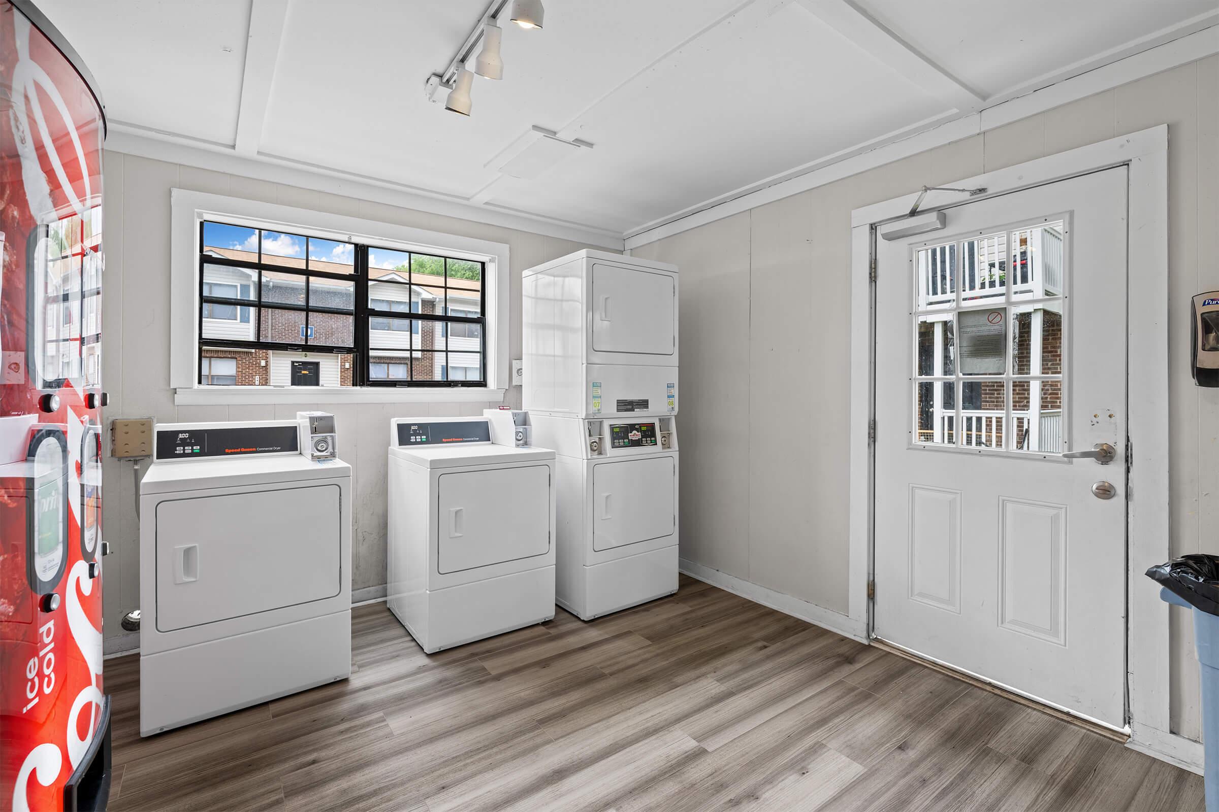 A clean and well-lit laundry room featuring three white washing machines and dryers side by side. A door leads outside, with a window showing an outside view of a building complex. The floor is covered in light-colored wood-like laminate, creating a bright and inviting atmosphere.