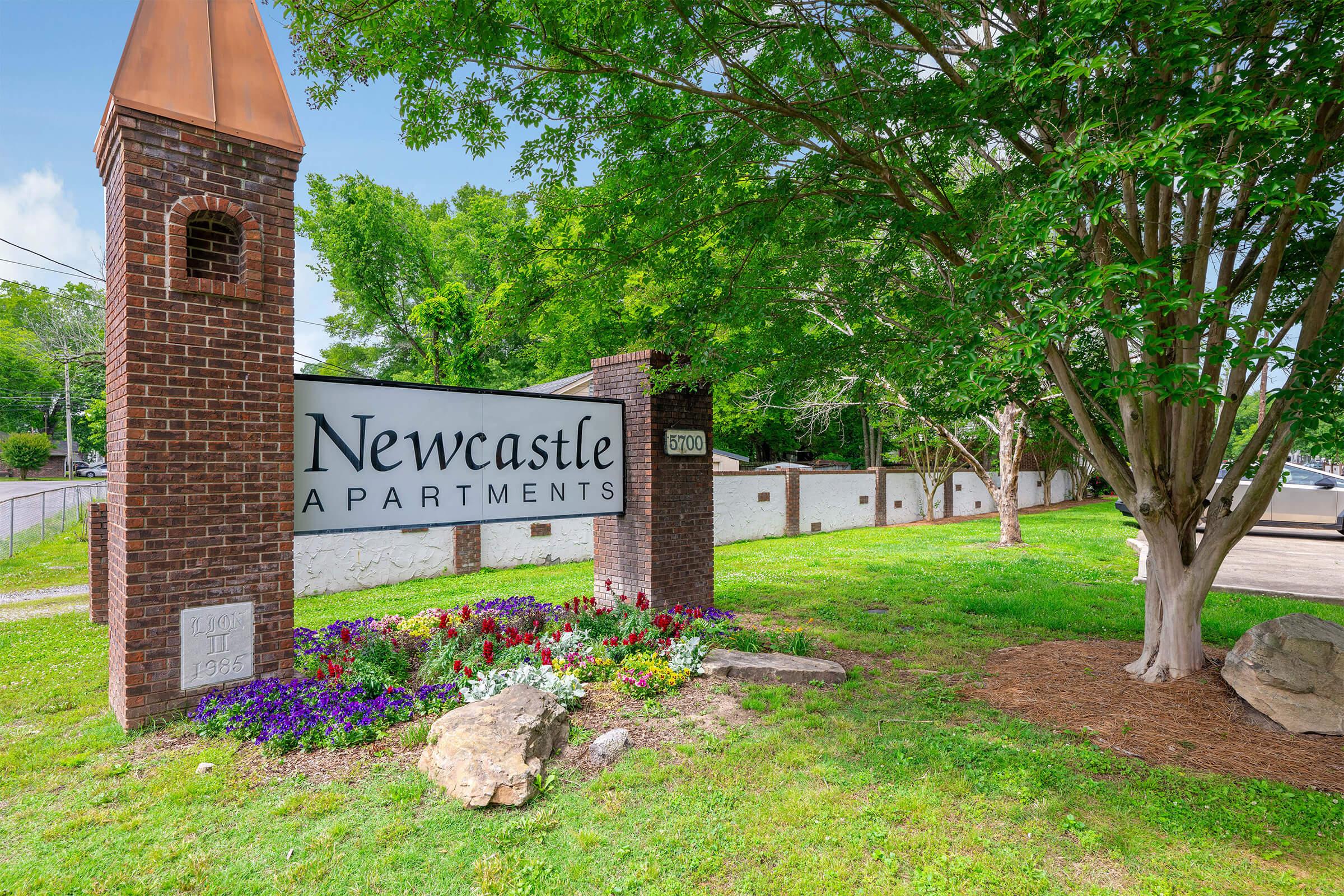 Sign for Newcastle Apartments, featuring brick columns, a copper-topped peak, and landscaped flowers in front. The setting includes green grass and trees, suggesting a welcoming residential environment. The sign displays "Newcastle Apartments" clearly for visibility.
