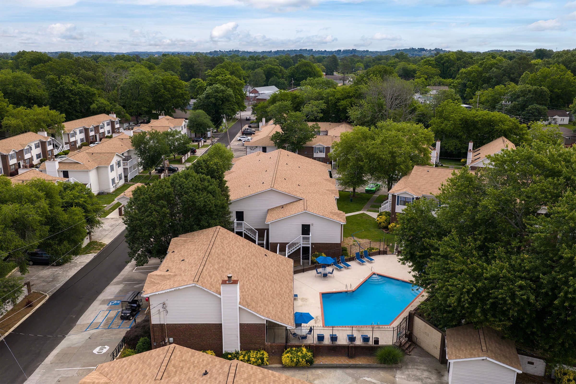 Aerial view of a residential area featuring several houses with brown roofs. In the center, there is a swimming pool surrounded by lounge chairs and green landscaping. The scene is set against a backdrop of trees and a clear blue sky.
