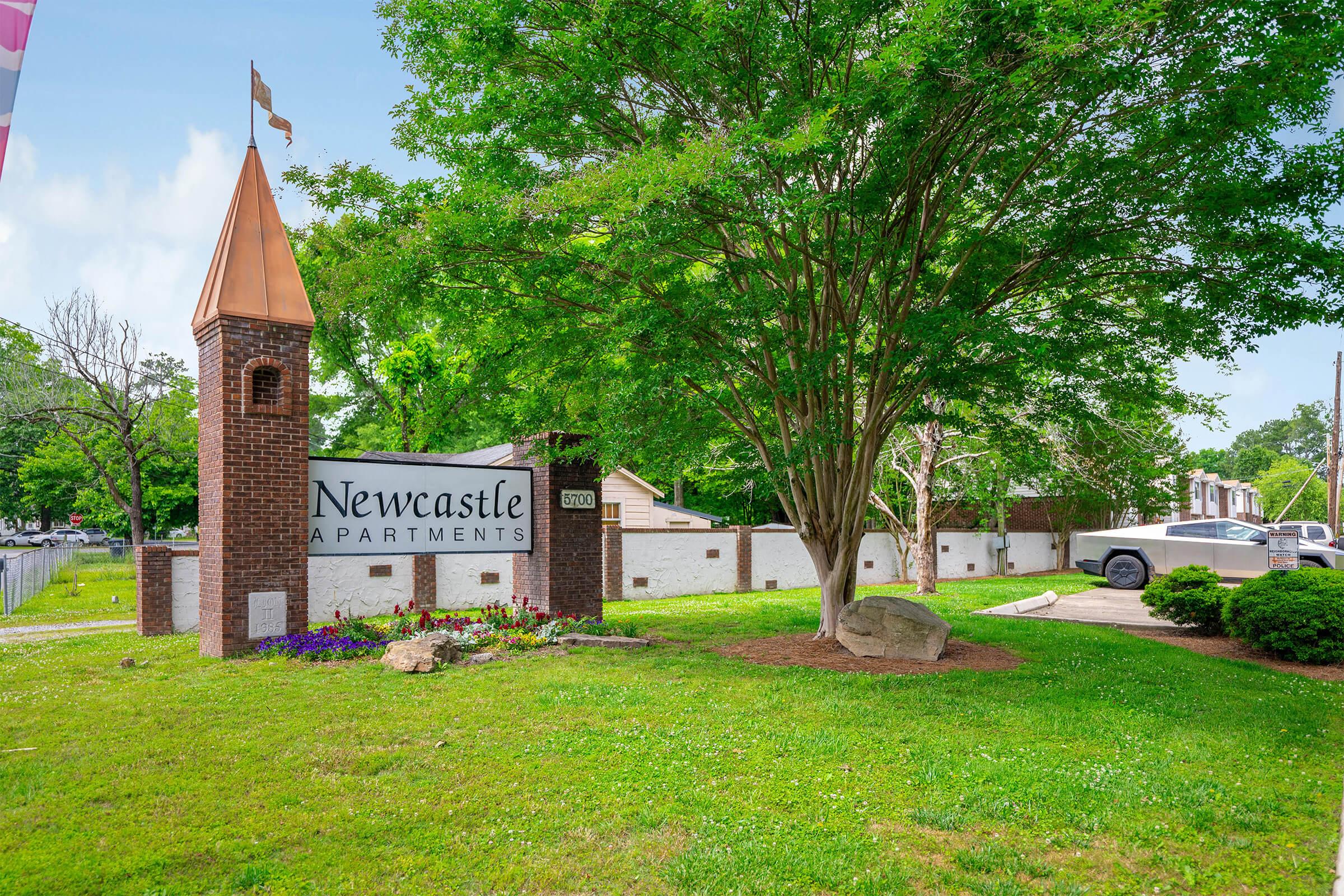Sign for Newcastle Apartments at the entrance, featuring a decorative tower with a flag. Surrounding greenery includes trees and flower beds, with a gravel driveway visible. A silver vehicle is parked nearby, and a white fence can be seen in the background. The scene conveys a welcoming residential atmosphere.
