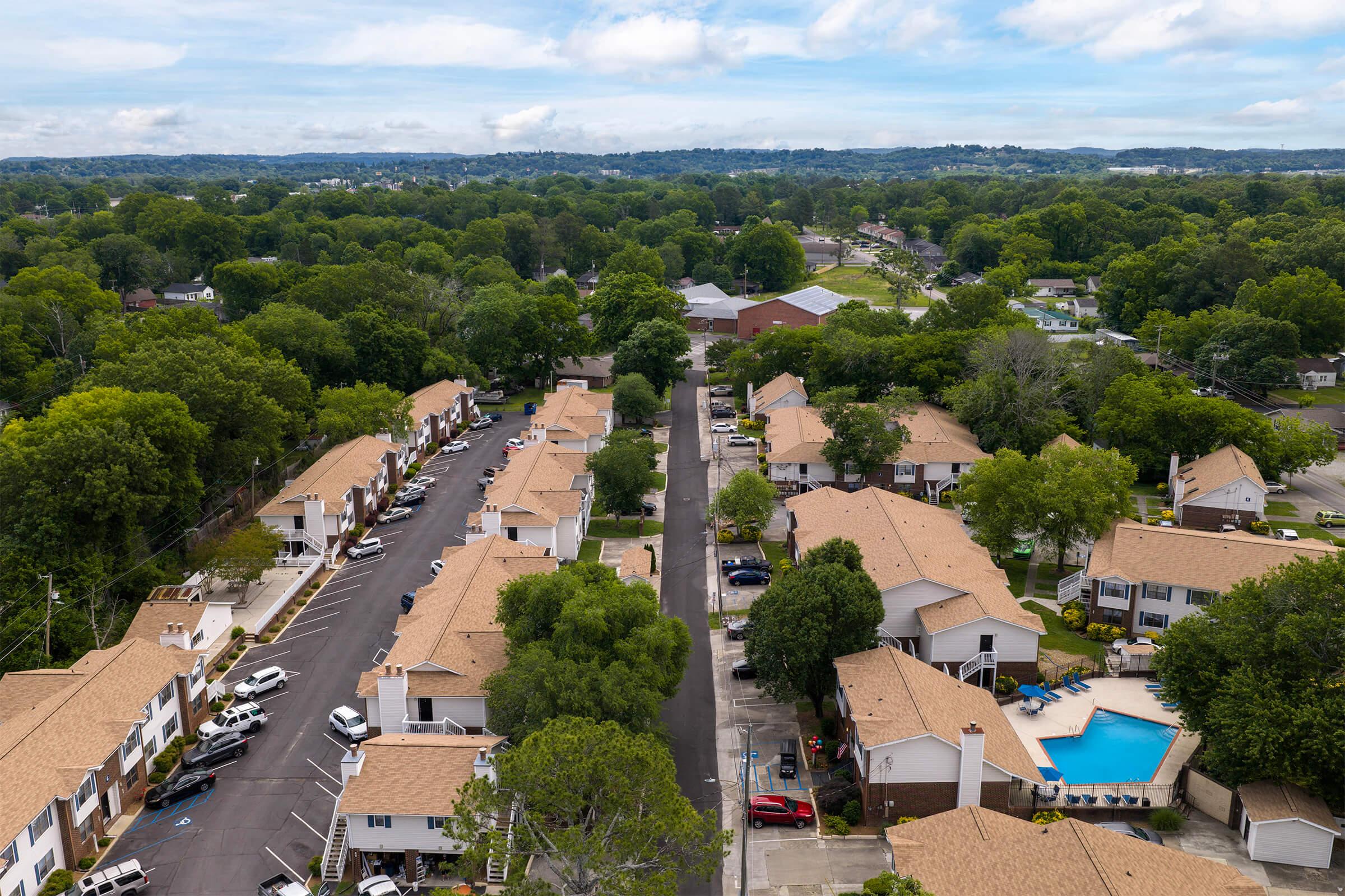 Aerial view of a suburban neighborhood featuring rows of single-story residential buildings with brown roofs. The scene is surrounded by lush green trees, and a clear blue sky with some clouds. A swimming pool is visible within the neighborhood, along with parked cars along the streets.