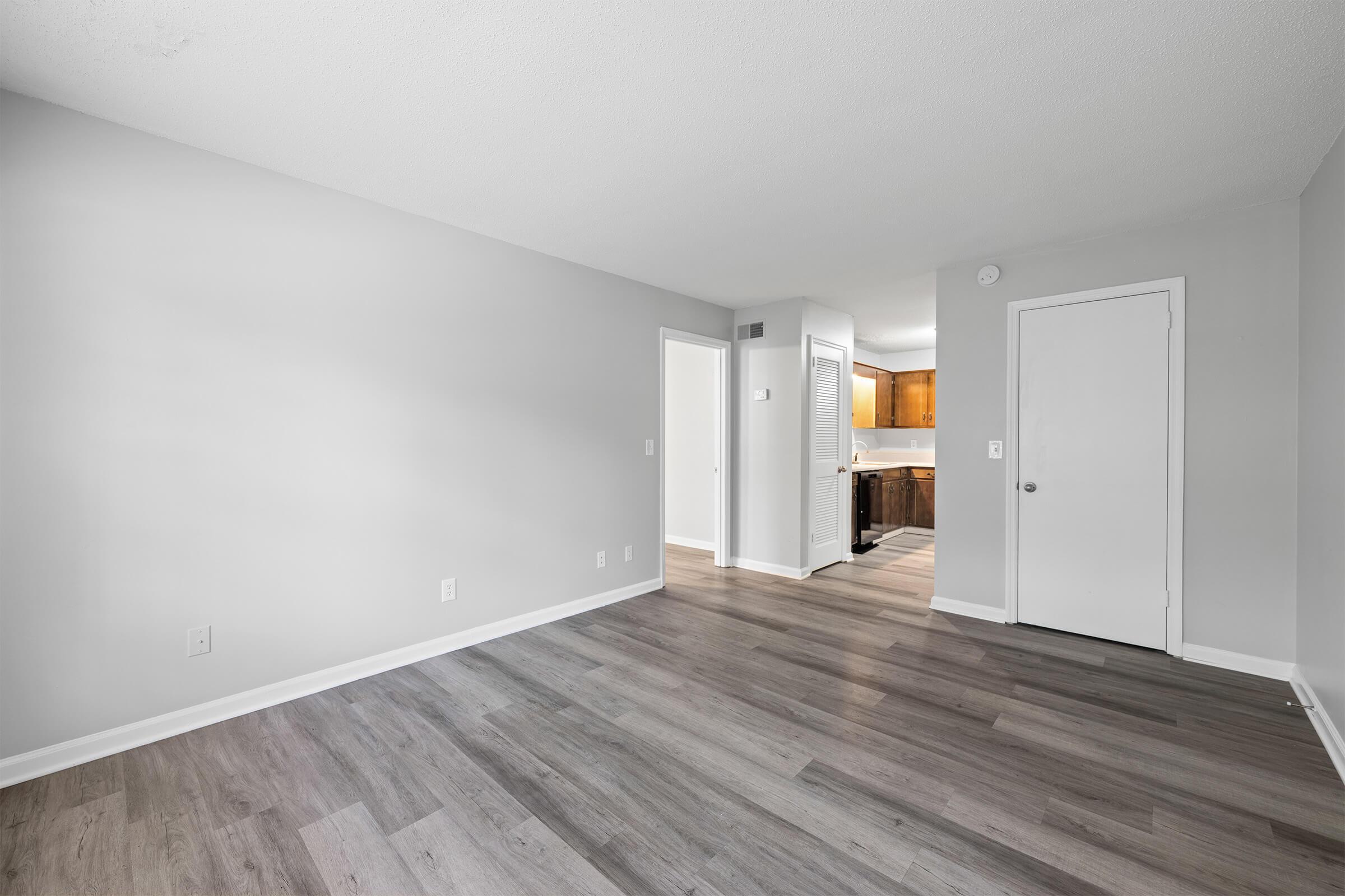 A spacious, empty room with light gray walls and dark wood-like flooring. The room features a doorway leading to a kitchen area with warm lighting. There's a closet door on the right and a plain white door on the far right. The overall atmosphere is bright and inviting, suitable for various decorating options.