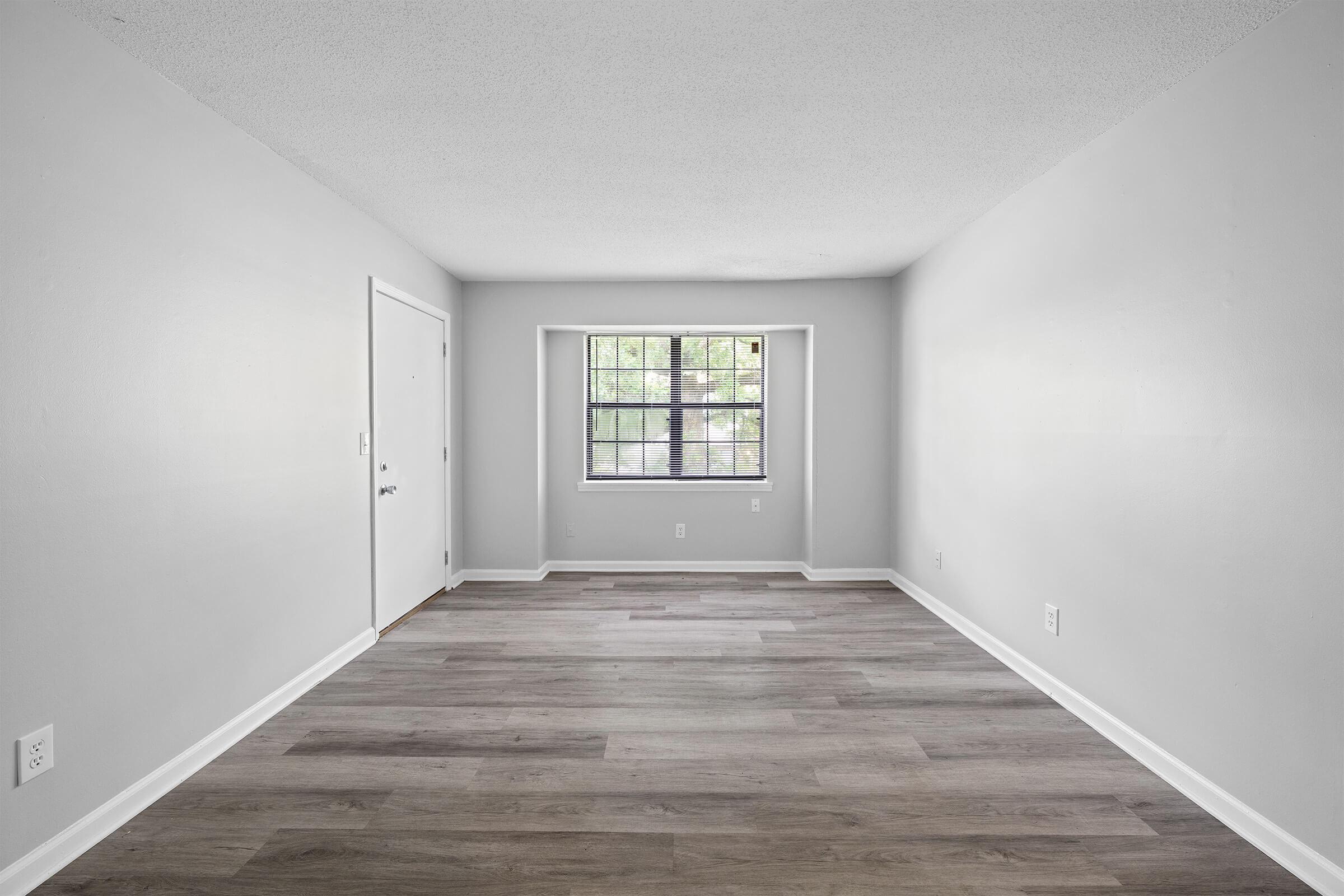 Empty room with light gray walls and a wood-style floor. A single window with a grid design lets in natural light, and a door is visible on the left. The space appears clean and minimalistic, suitable for various uses or furnishings. No furniture or decor is present.