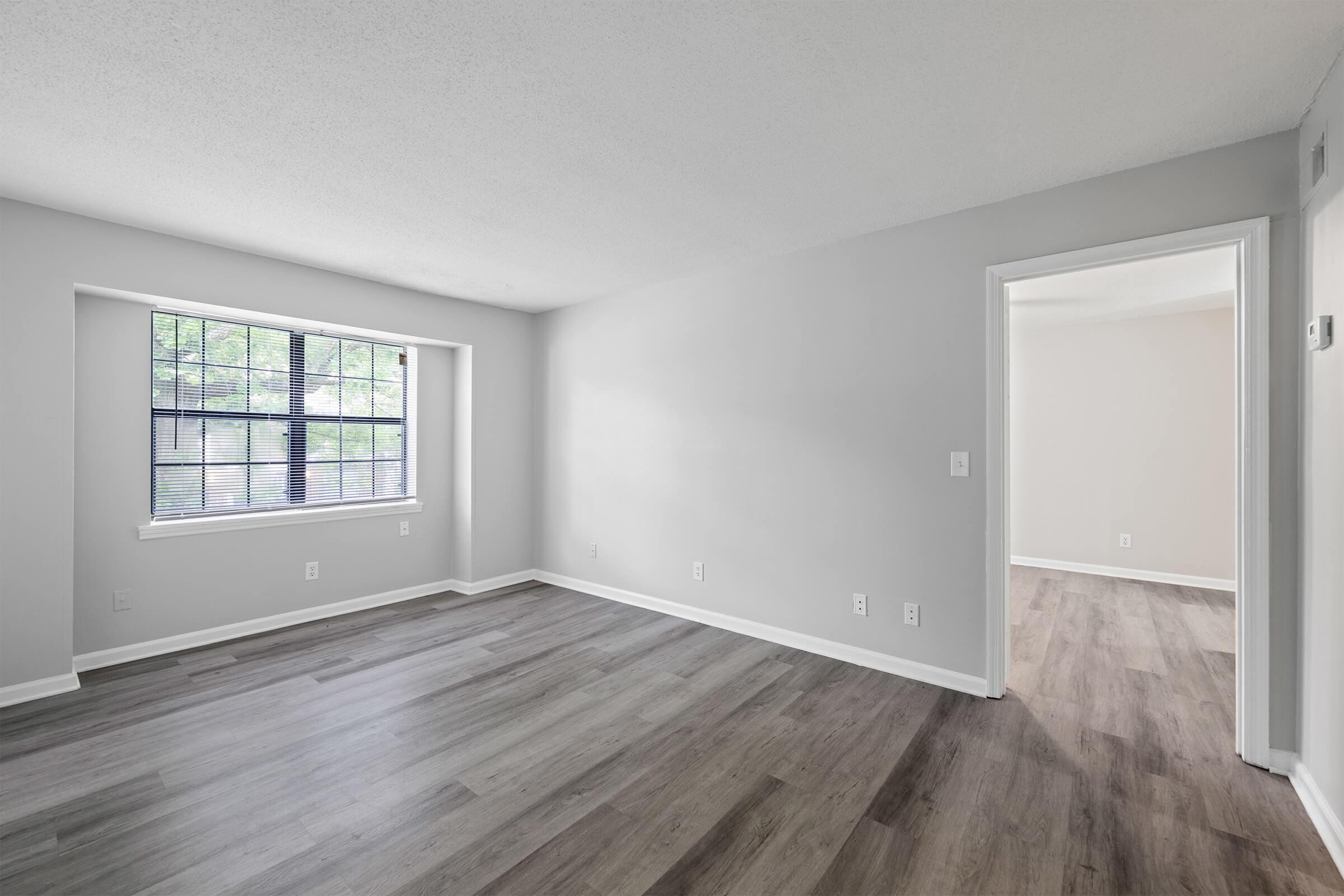 A bright, empty room featuring light gray walls and wood-like flooring. A window with black-framed panes is visible on the left side, allowing natural light to enter. An open doorway on the right leads to another light-colored room, suggesting a spacious layout.
