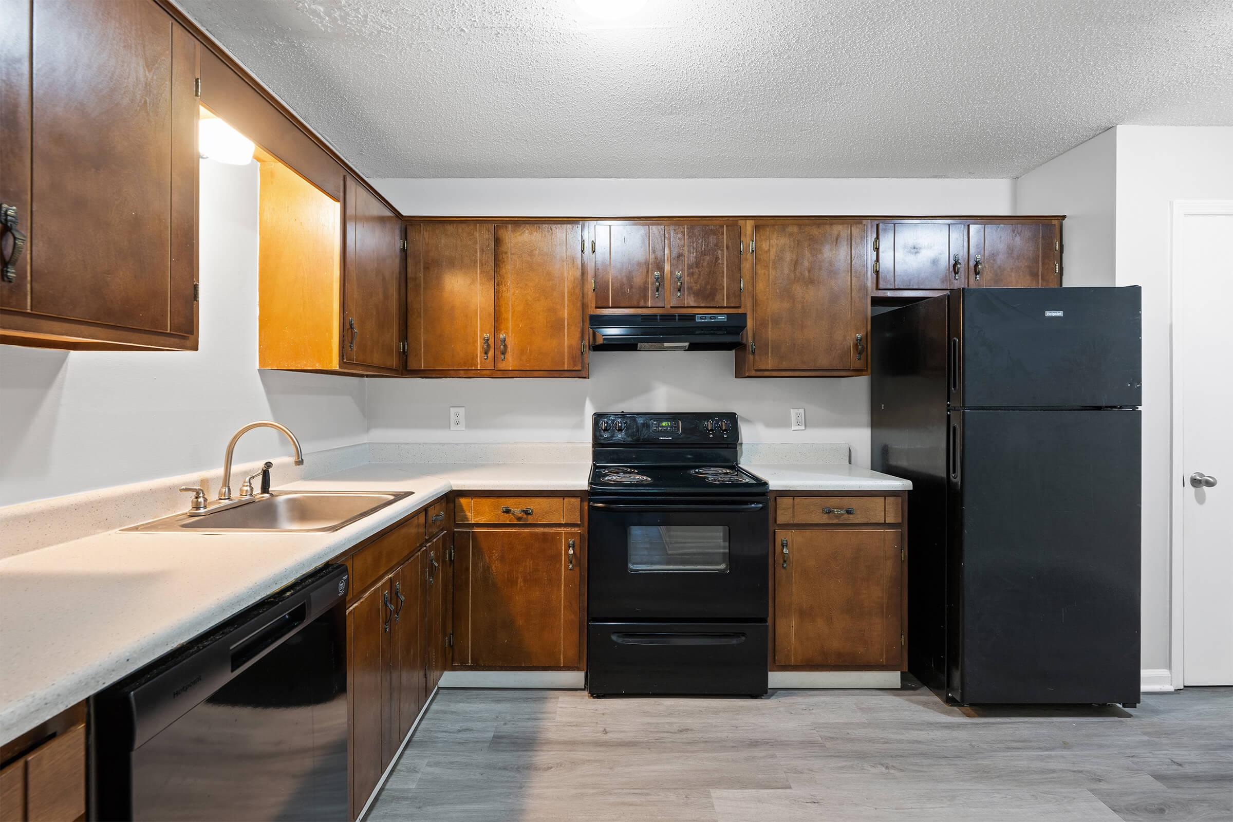 A modern kitchen featuring wooden cabinets, a stainless steel sink, and black appliances, including an oven and refrigerator. The countertop is light-colored, and the flooring is a light laminate. The space is well-lit with overhead lighting.