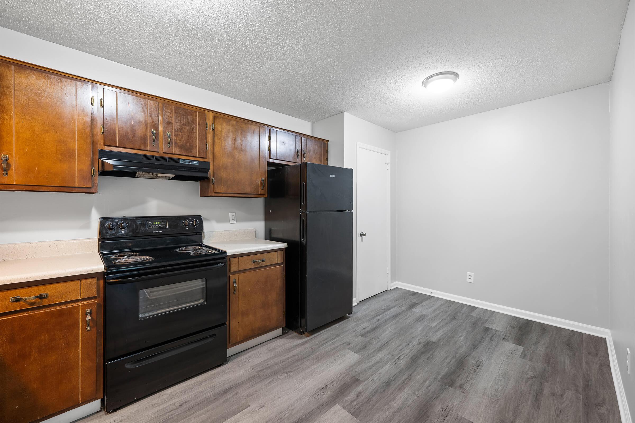 A modern kitchen featuring dark wooden cabinets, a black stove and refrigerator, a light-colored countertop, and gray flooring. The walls are painted in a light gray, and there is a door leading to another room visible in the background.