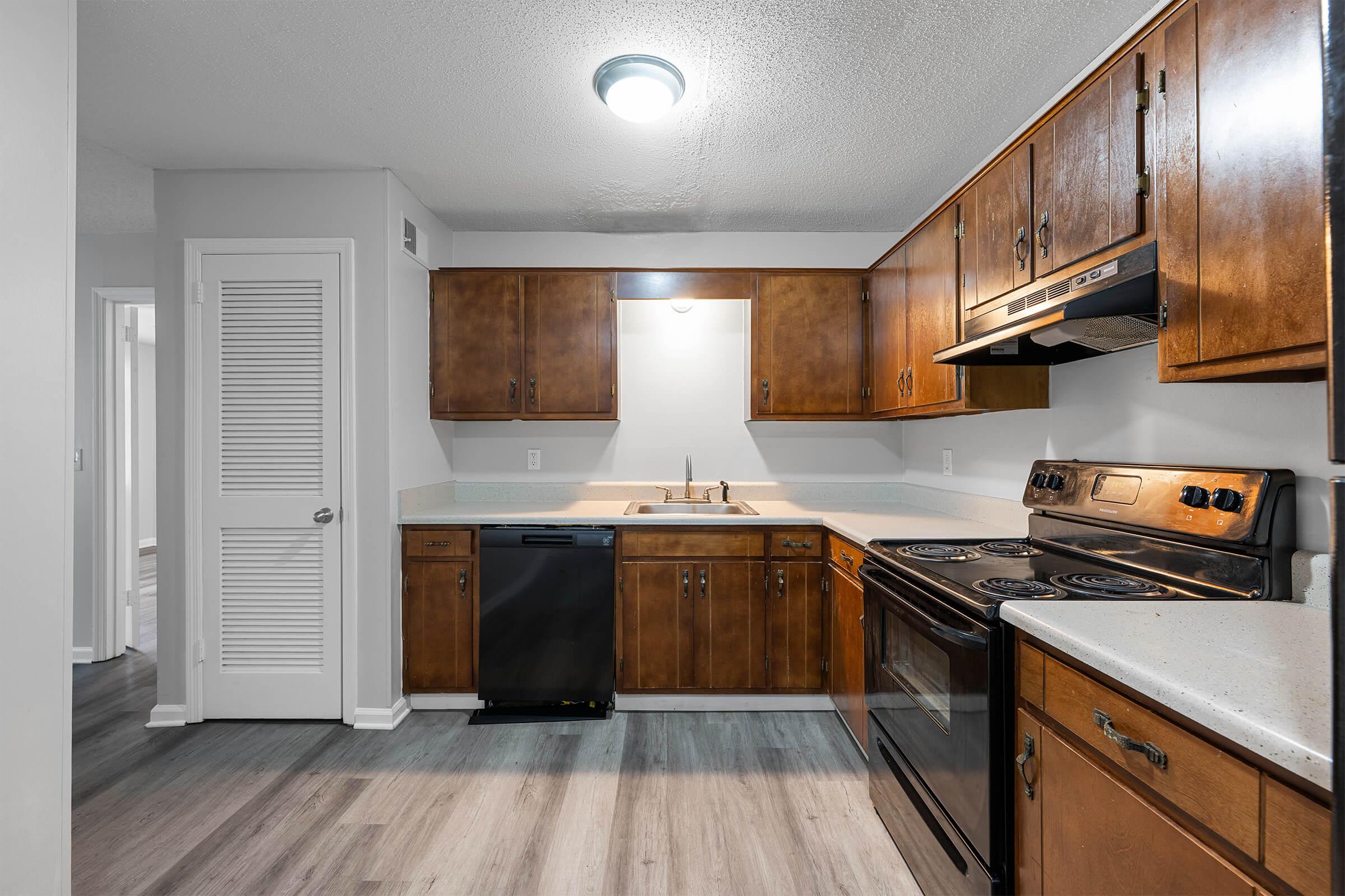 A modern kitchen featuring wooden cabinets, a stainless steel sink, and black appliances including a dishwasher and stove. The countertops are light-colored, and the flooring is a wood-like laminate. The kitchen is well-lit with a ceiling light and has a neutral color palette.