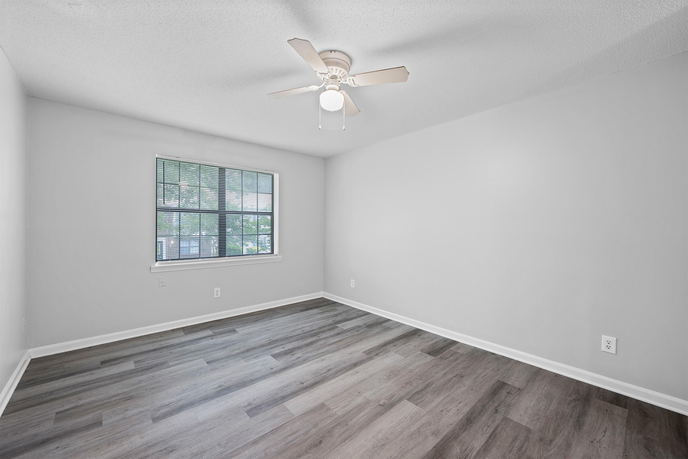 A spacious, empty room with light gray walls and light wood laminate flooring. A ceiling fan is mounted in the center, and a window with blinds lets in natural light, offering a view of greenery outside. The overall atmosphere is bright and clean, suitable for various decorative styles.