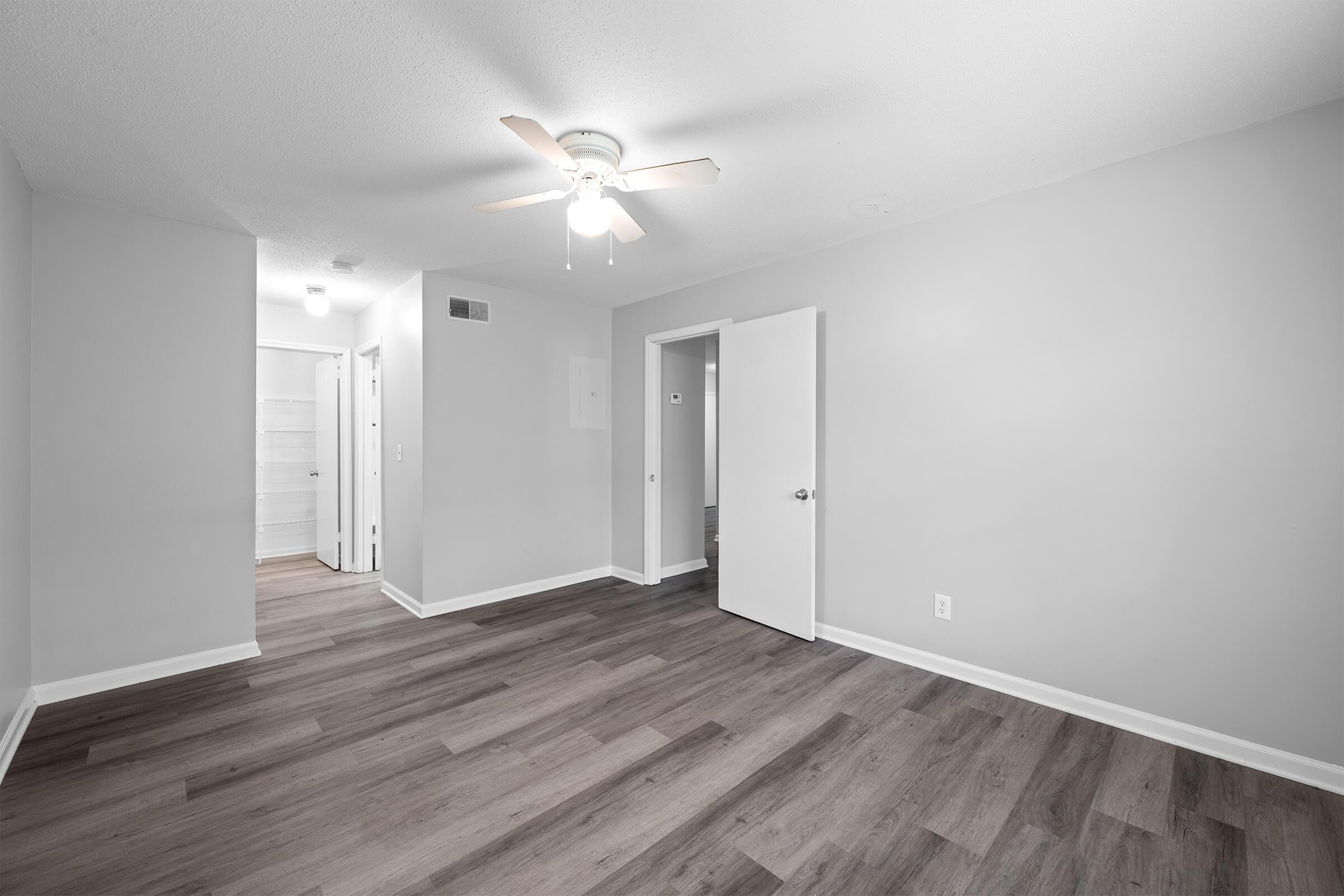 An interior view of a freshly painted room featuring light gray walls and wood-style flooring. The room includes a ceiling fan, a doorway leading to a hallway, and doors to other rooms. Natural light enters from an unseen source, creating a bright and open atmosphere.