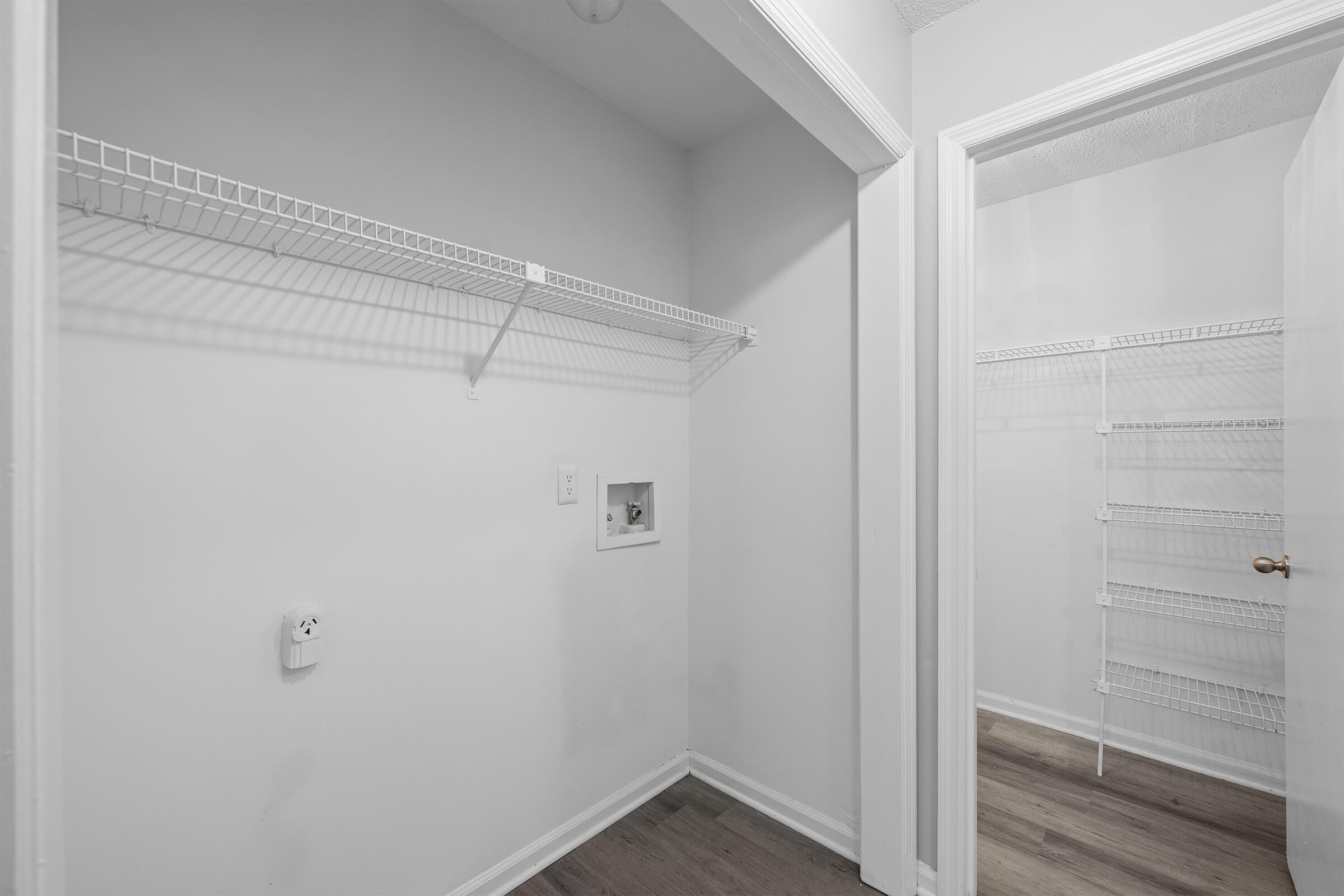 A small, empty room featuring white walls and wooden flooring. On the left, there's a wire shelf against the wall, and a small electrical outlet is visible. To the right, an open door leads to another room with additional wire shelving, indicating the space could be used for storage.