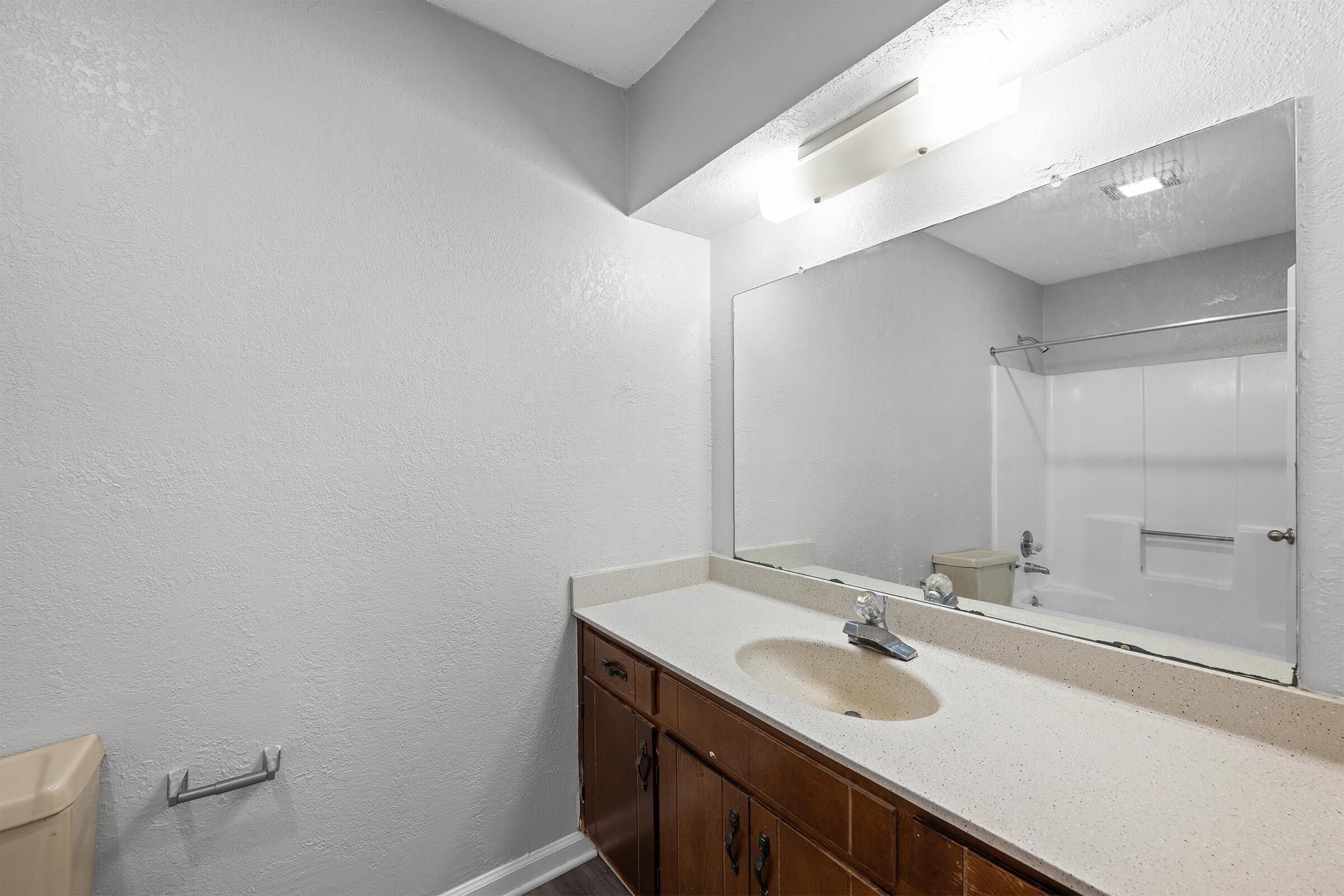 Modern bathroom with a light gray textured wall, a large mirror above a beige countertop with a single faucet, and wooden cabinetry. Visible features include a white bathtub in the background, a toilet on the left, and a towel holder by the sink. Soft overhead lighting illuminates the space.