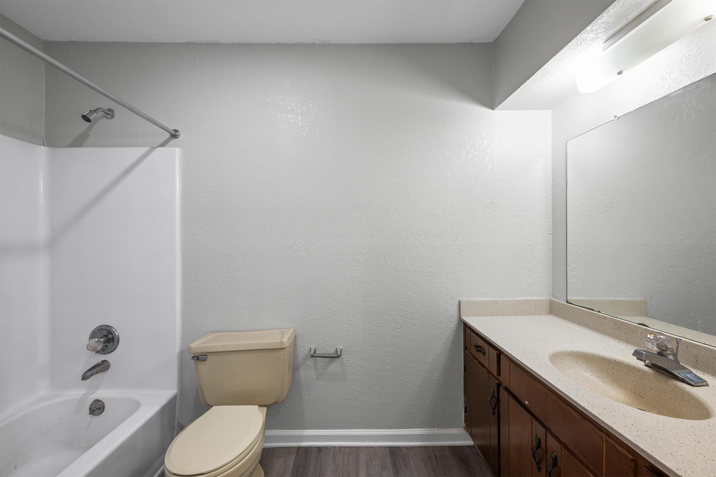 A clean and simple bathroom featuring a bathtub with a shower, a beige toilet, and a single sink vanity with a light-colored countertop. The walls are painted gray, and there is a large mirror above the sink. The flooring appears to be laminate in a wood-like finish.