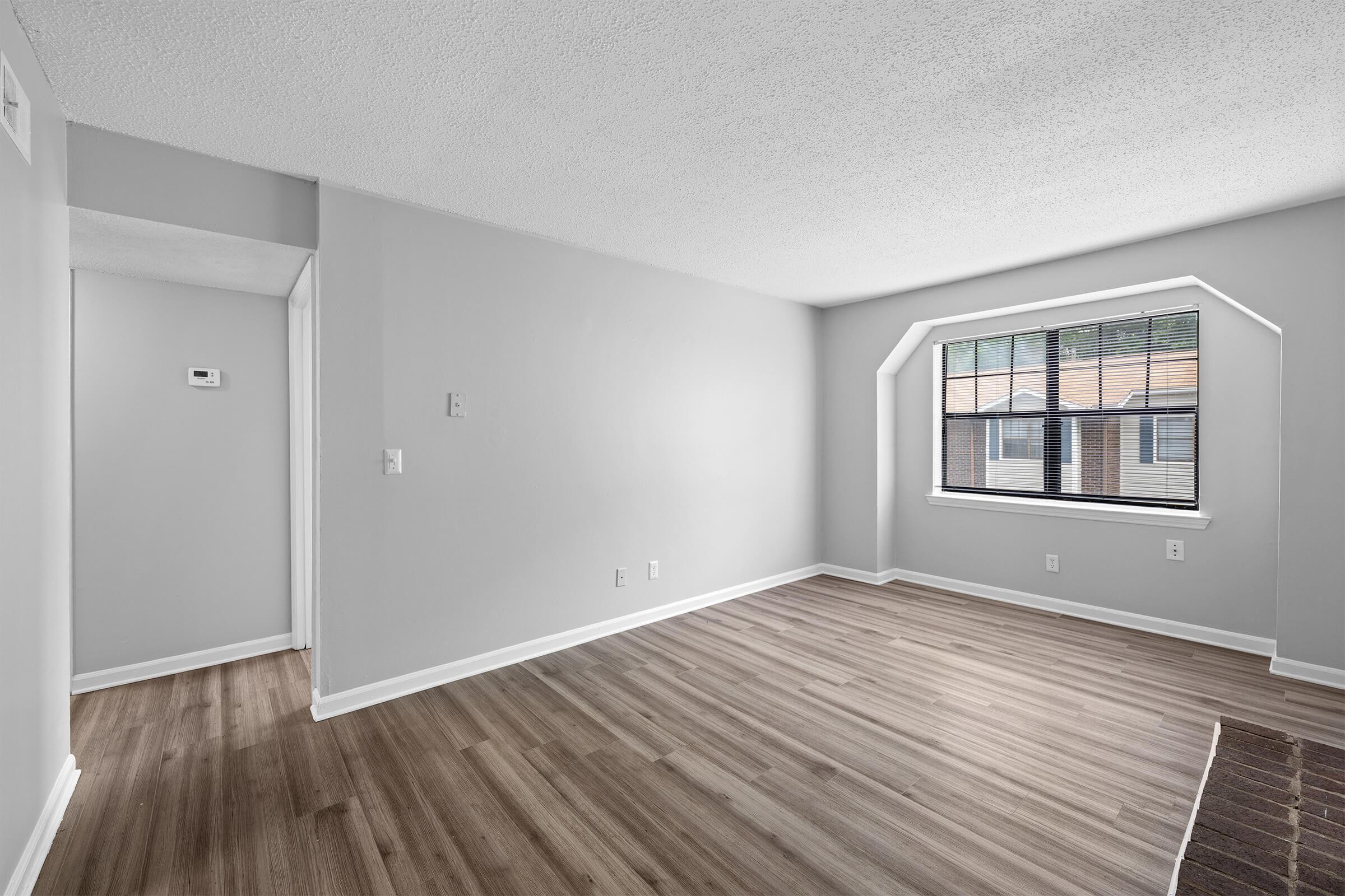 A spacious, empty living room featuring light gray walls and laminate flooring. A large bay window lets in natural light, and there's a small hallway visible to the left. The room is ready for furnishings, creating a blank canvas for decoration or design.