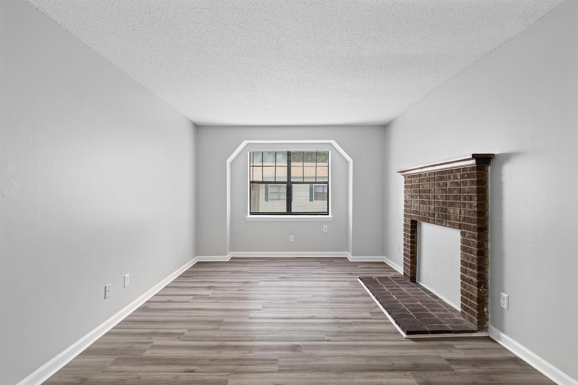 Empty living room with light gray walls and wood-like flooring. A large window with a grid pattern provides natural light. There is a decorative brick fireplace along one wall, featuring a simple mantel and dark tiles at the base, creating a spacious and airy feel.