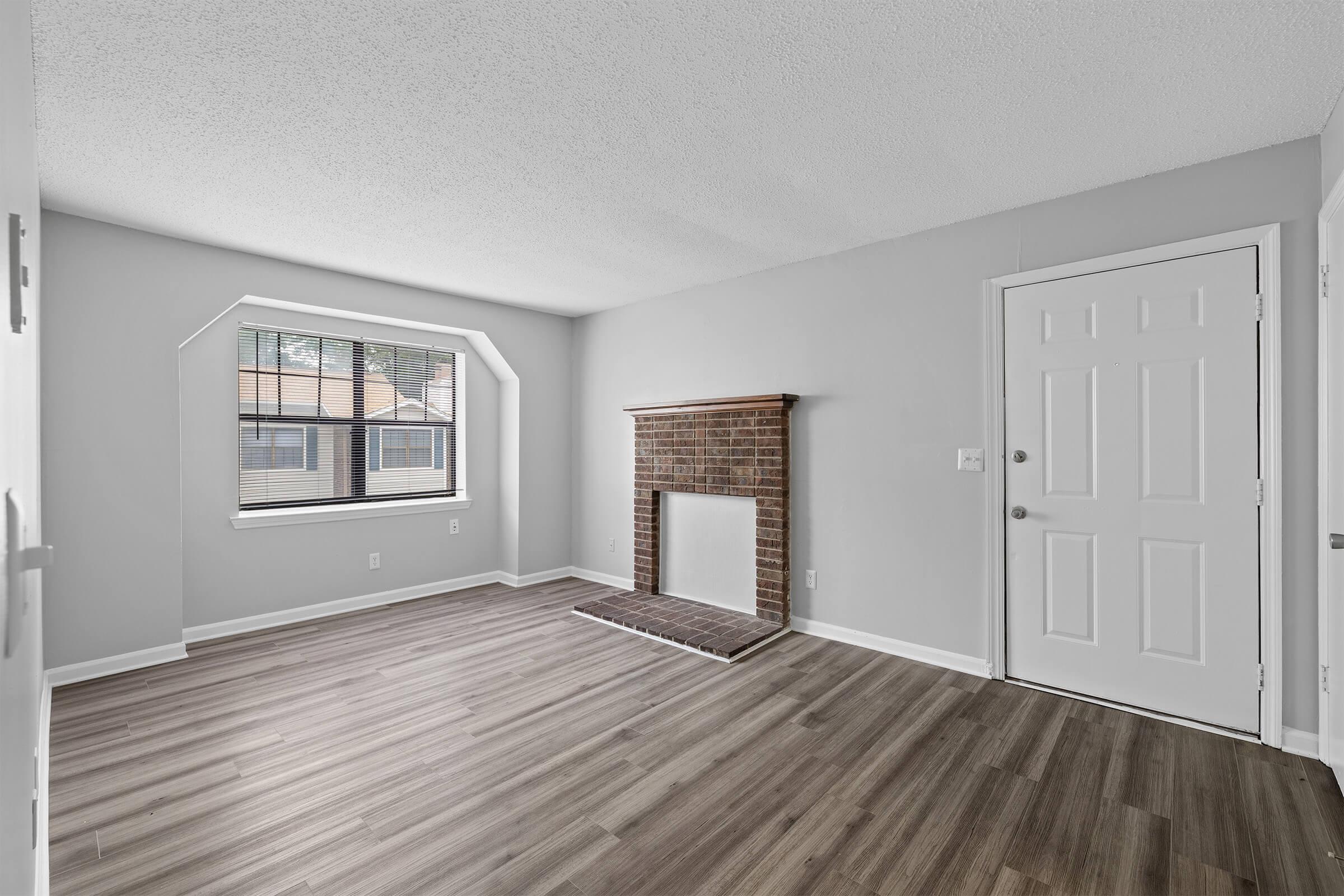 A spacious living room featuring light gray walls, a large window with a unique arch design, and a brick fireplace as a focal point. The floor is covered with wood-like laminate, and a white front door is visible on the right. Natural light fills the room, creating an inviting atmosphere.