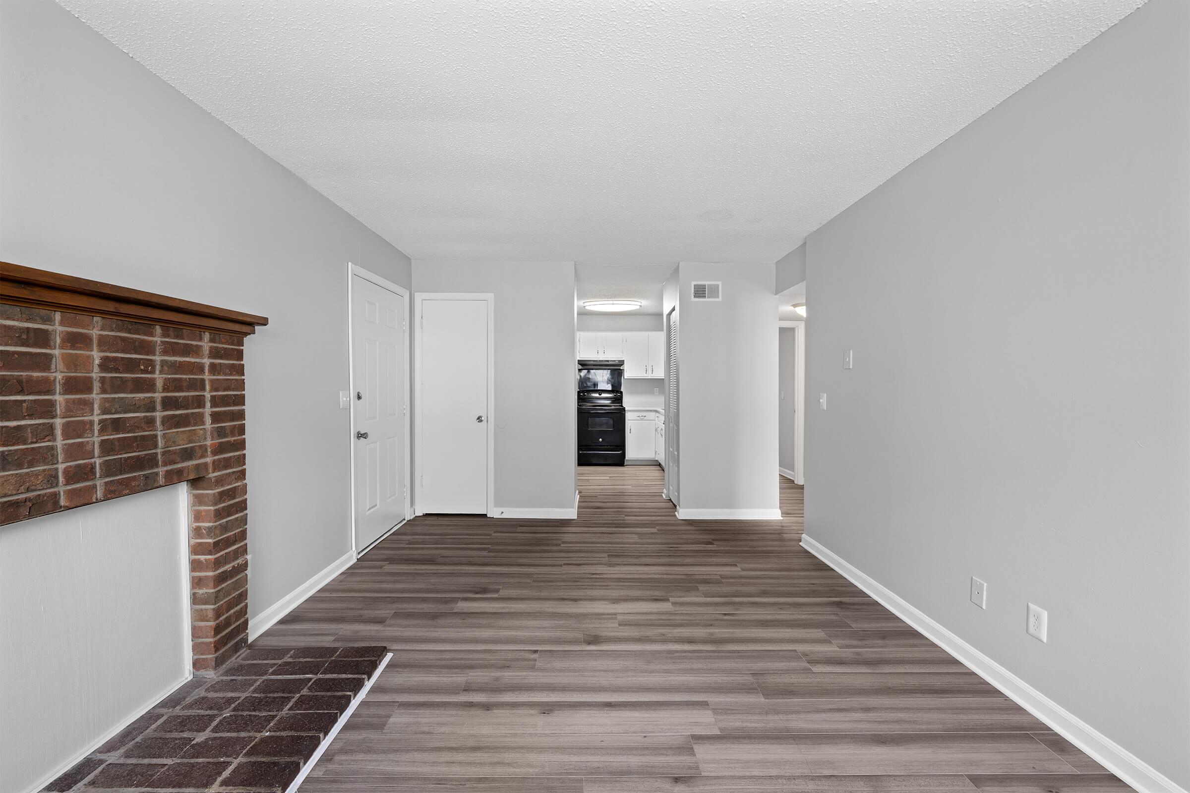 A spacious, well-lit living area featuring light gray walls and wooden flooring. A brick fireplace is on the left, and a door leads to another room. In the background, a doorway opens into a kitchen area with modern appliances. The overall atmosphere is clean and modern, suitable for comfortable living.