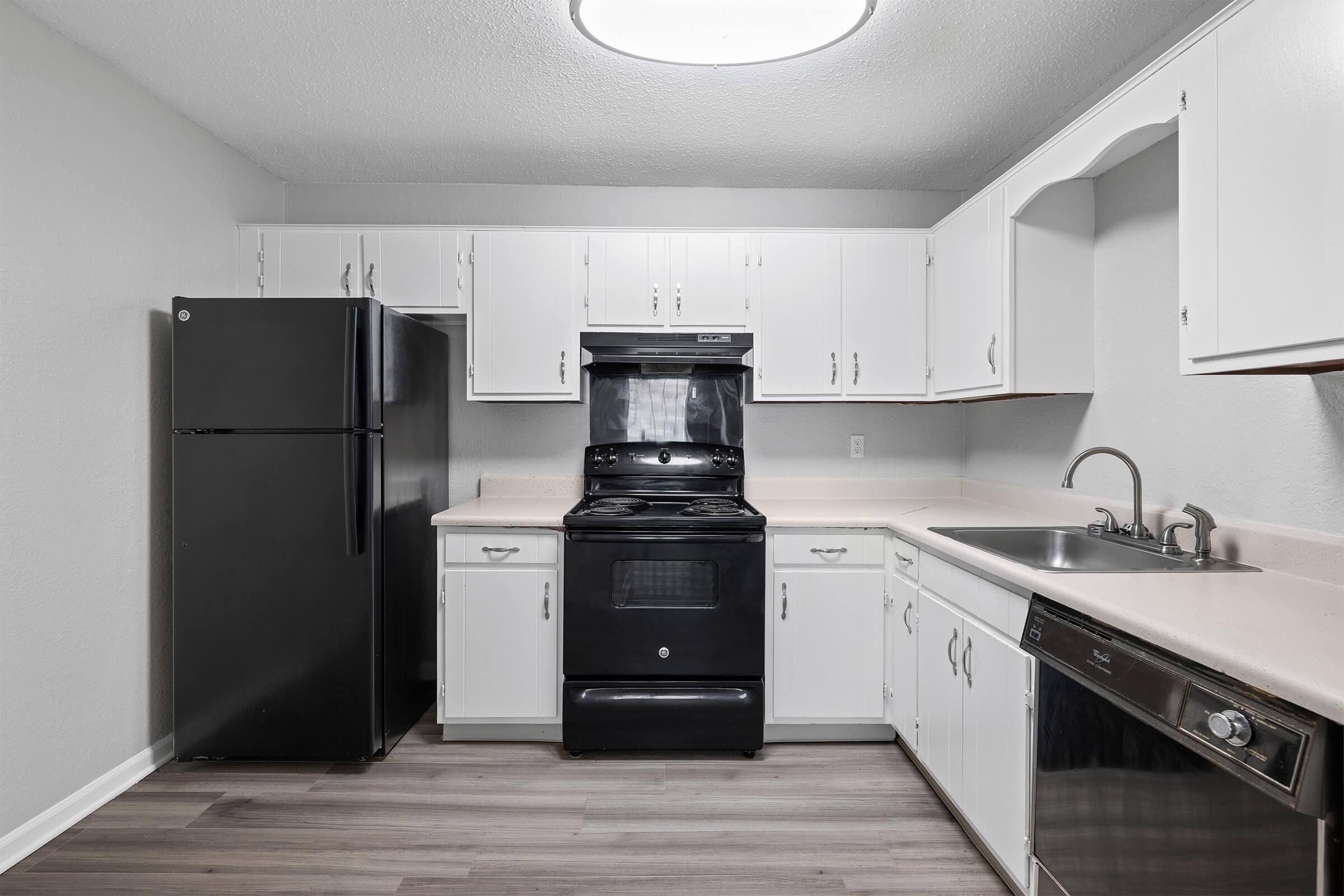 A modern kitchen featuring white cabinets, a black refrigerator, and a black oven with a stovetop. There is a stainless steel sink with a single faucet and a dishwasher. The countertops are light-colored, and the flooring appears to be laminate. The walls are painted a light gray.