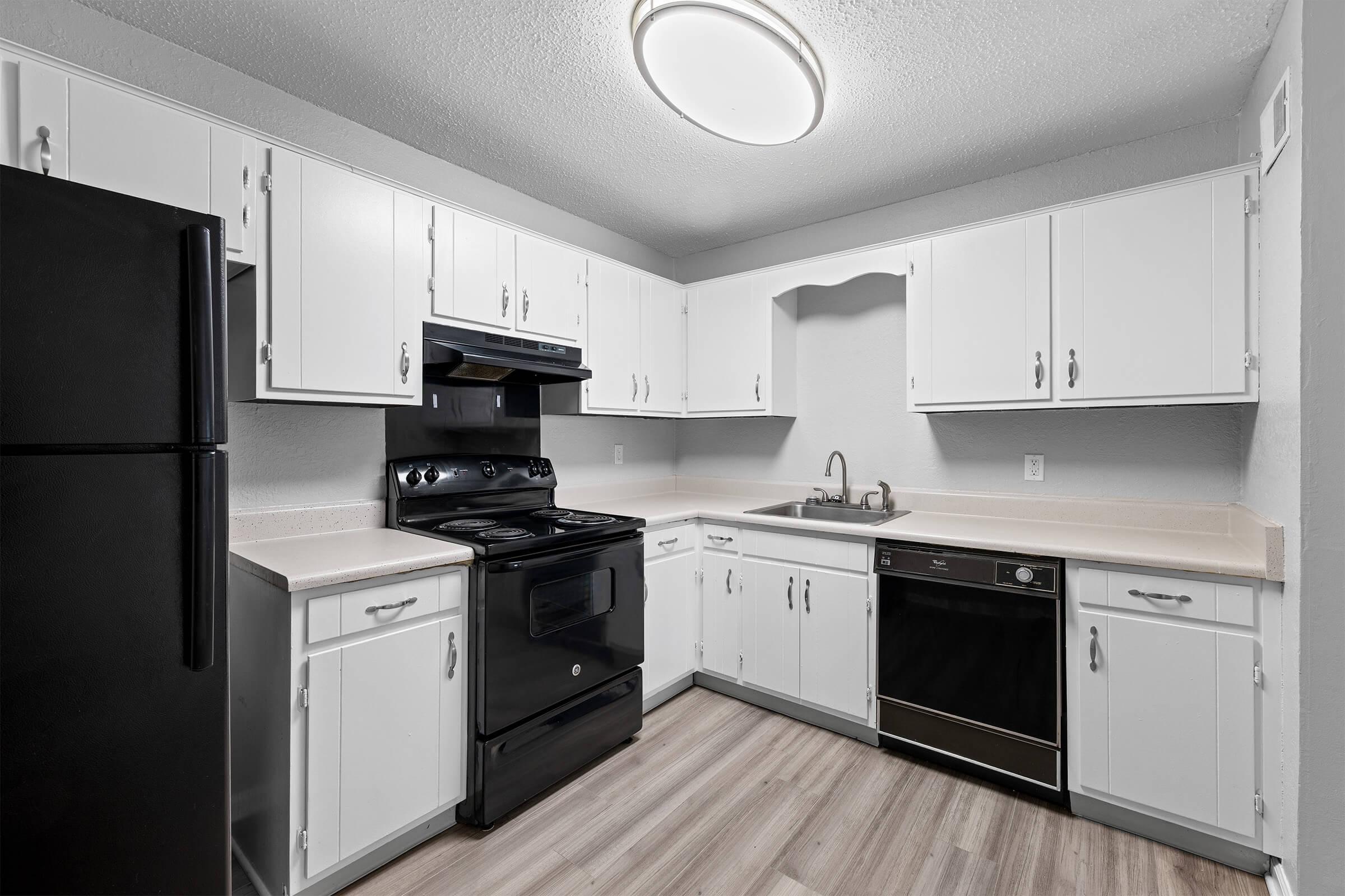 A modern kitchen featuring white cabinetry, a black refrigerator, and a black stove. The countertops are light-colored, and there is a stainless steel sink and dishwasher. The flooring is a light wood laminate, and the space is illuminated by a circular ceiling light.