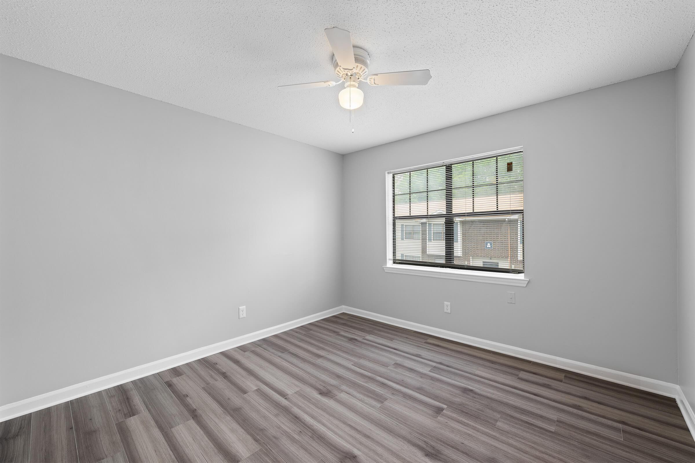 A spacious, empty room with light gray walls and a ceiling fan. The flooring is wood-like laminate in shades of gray. A large window with blinds allows natural light to enter, creating a bright ambiance. The room is ready for personalization and furnishing.