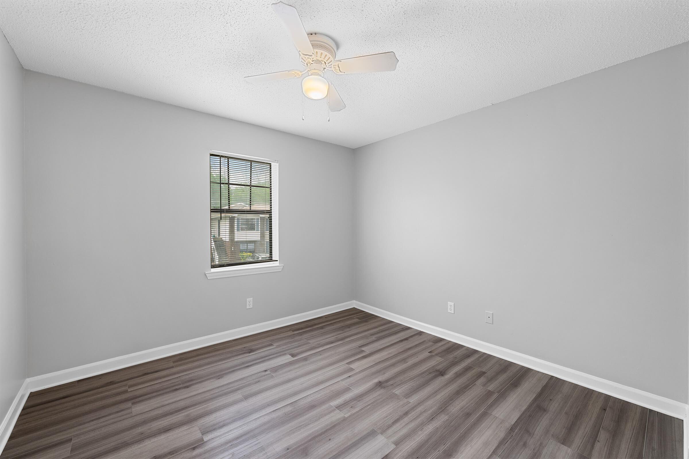 A small, empty room with gray walls and a ceiling fan. The floor is made of light-colored wood laminate. A single window with blinds allows natural light to enter, illuminating the space. The room has a minimalist design, making it suitable for various uses such as a bedroom or office.