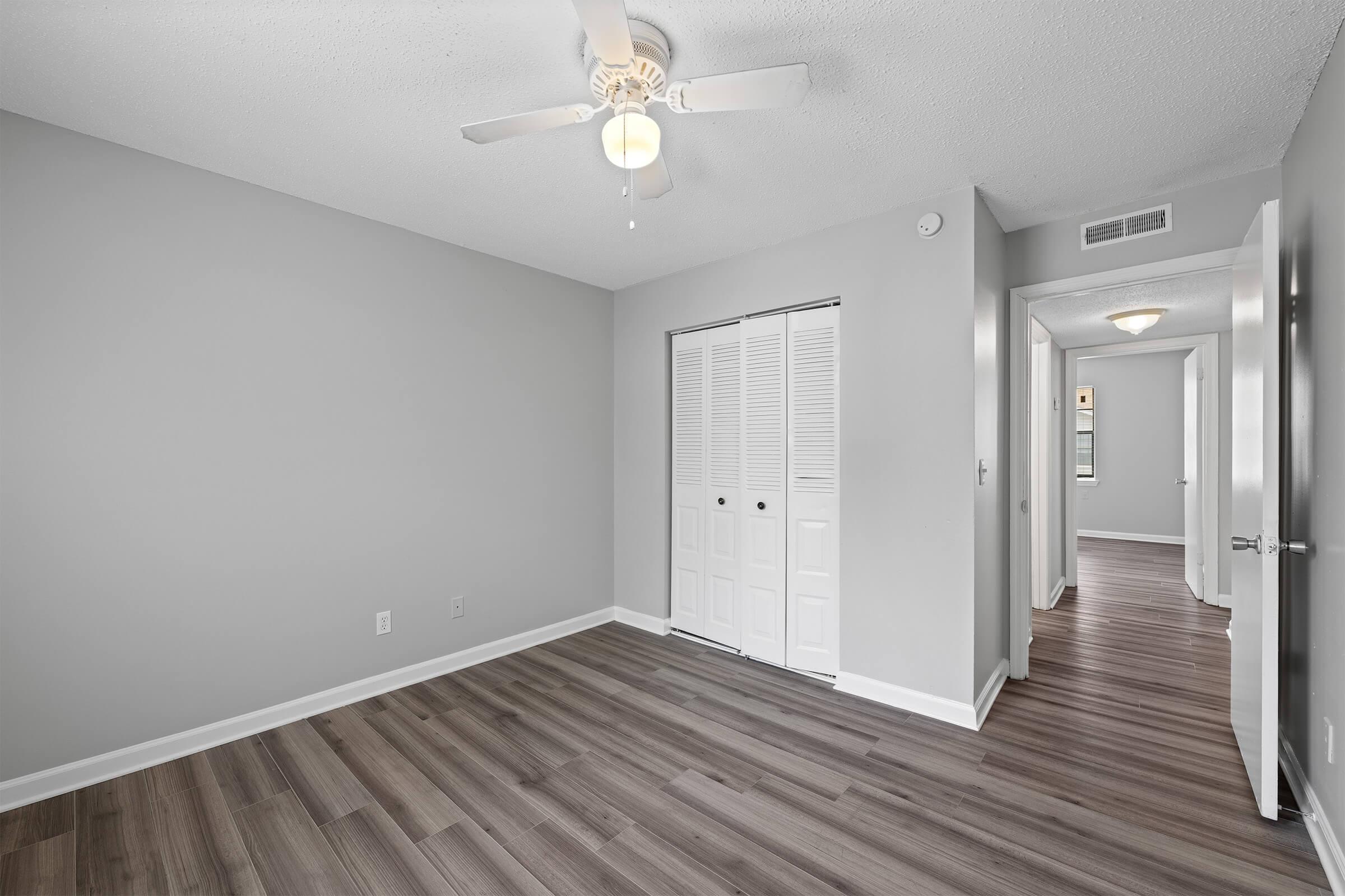 A spacious, unoccupied room with light gray walls and dark wood laminate flooring. A ceiling fan hangs from the ceiling, and there are two white closet doors. A hallway leads to another room in the background, with a door partially visible. Natural light comes from windows not shown in the image.