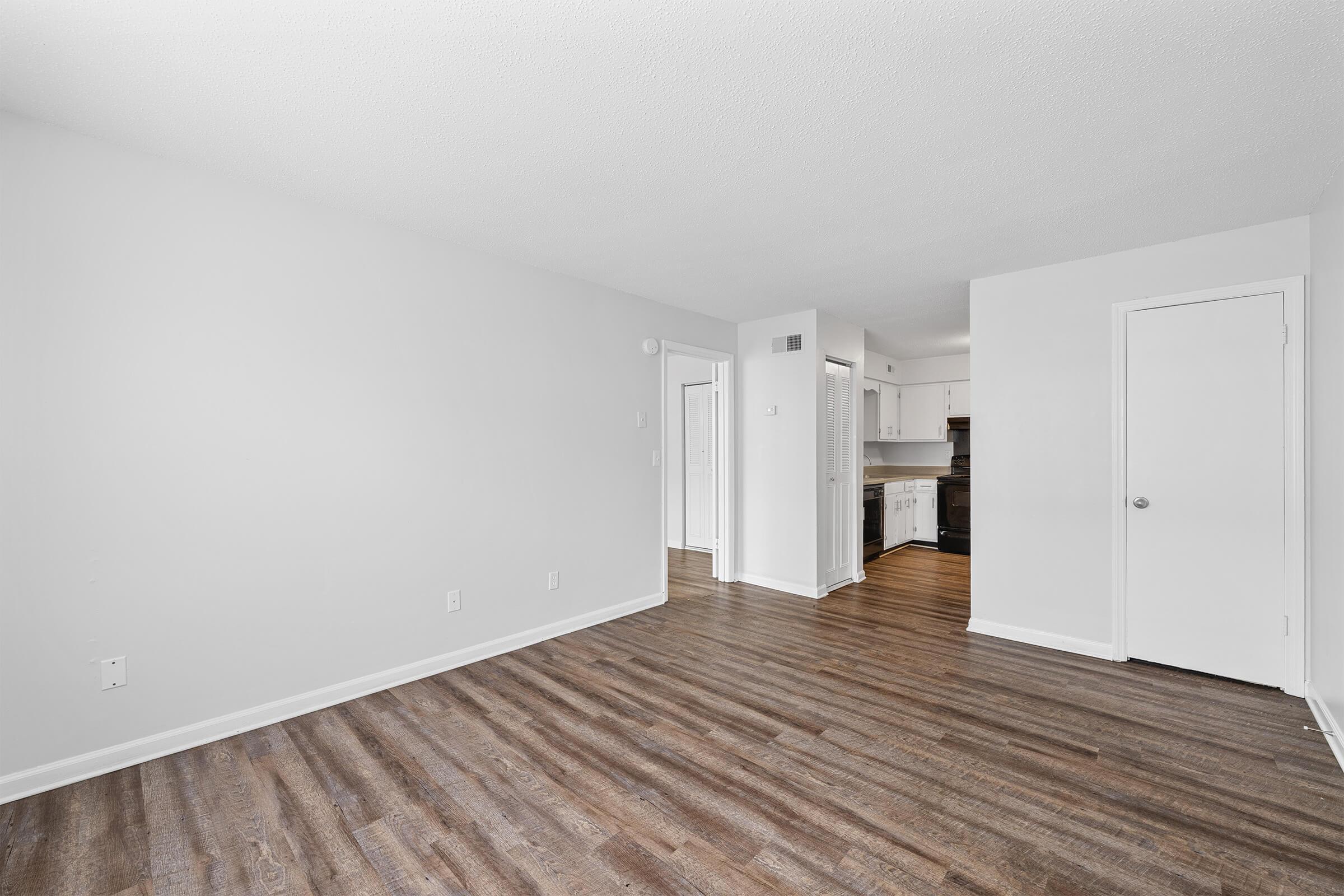 A bright, empty room with light gray walls and dark wood-like flooring. A doorway leads to a kitchen area in the background, and another door is visible on the right. The space is well-lit and spacious, showcasing a clean and modern design.