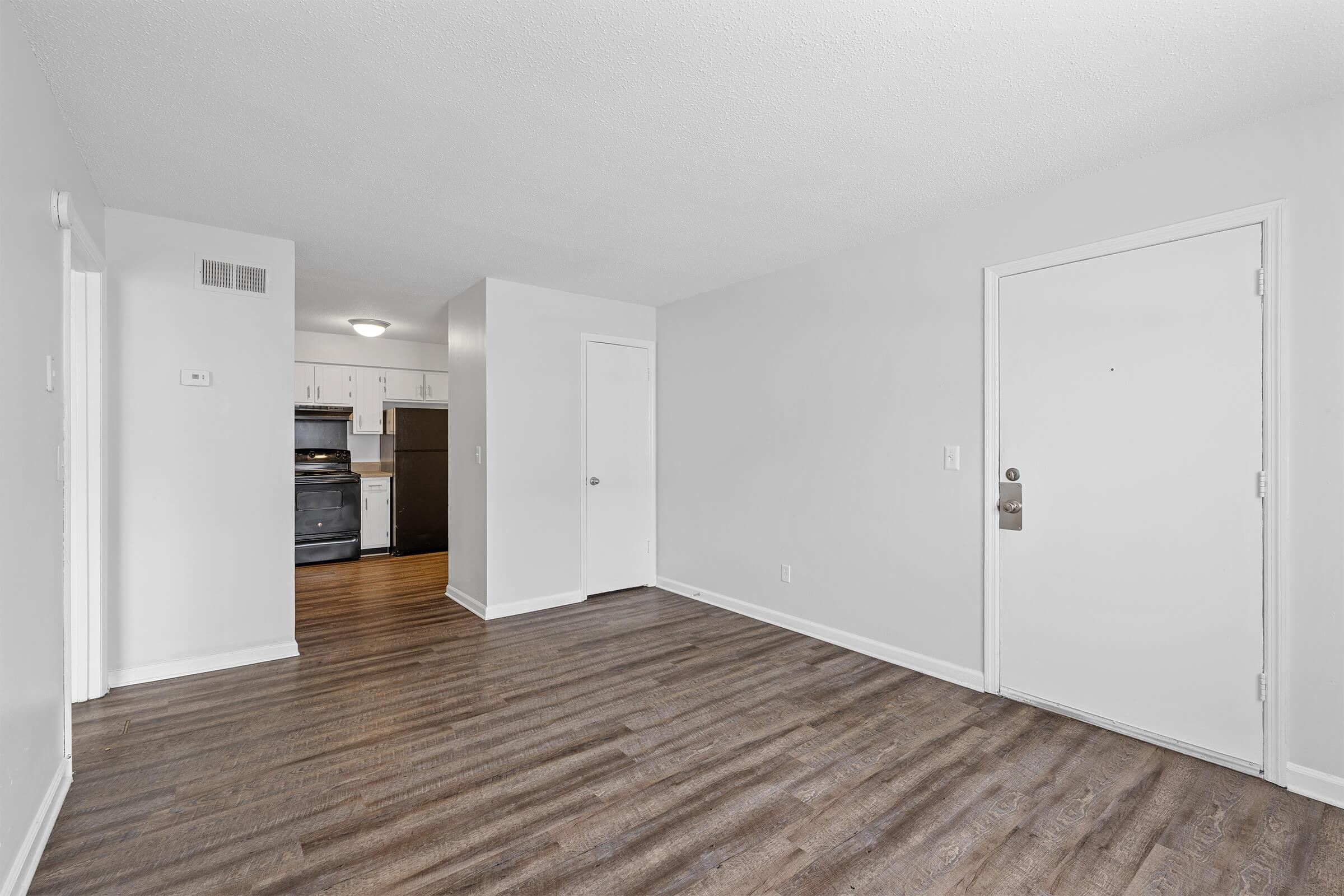 A bright, empty room with hardwood-like flooring and white walls. A door leads to another area, likely a kitchen, featuring appliances in the background. The main entrance is visible on the right side of the image, showcasing a simple design with a doorknob and a light switch on the wall.
