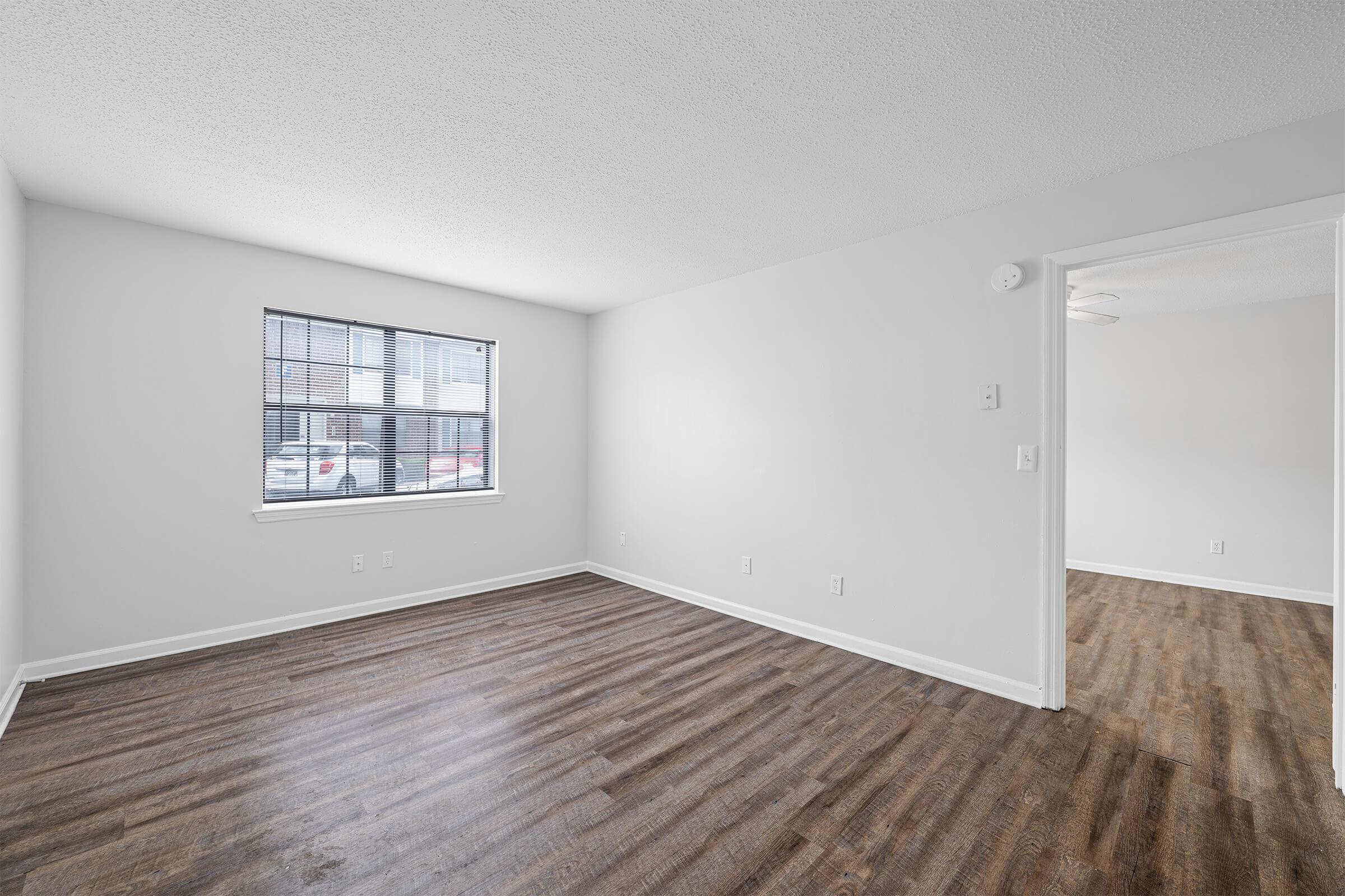 A spacious, empty room with light-colored walls and a large window featuring blinds. The floor is covered with dark wood laminate, and there is a doorway leading to another room visible in the background. The overall ambiance is bright and clean, suitable for various uses.