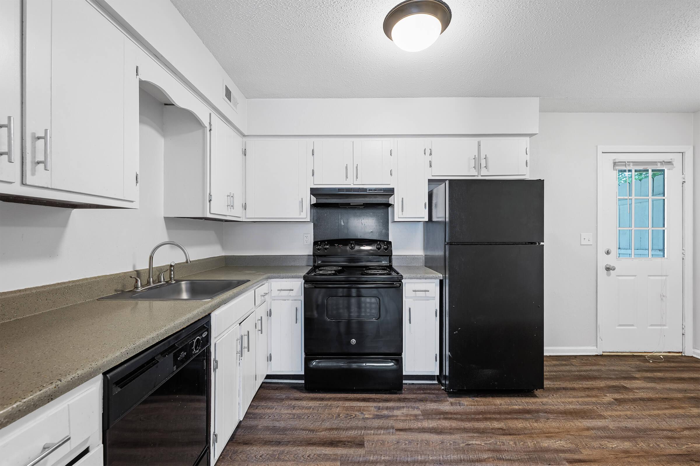 A modern kitchen featuring white cabinets, a black stove and refrigerator, a stainless steel sink, and dark wood laminate flooring. A door leads to the outside, and there is a window above the sink allowing natural light into the space.