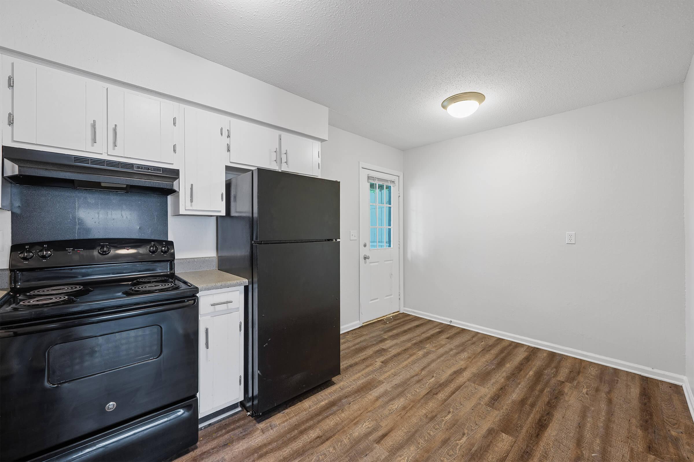 A view of a kitchen featuring black appliances, including a stove and refrigerator, with white cabinets. The flooring is wood-like, and there is a door leading outside. The walls are painted white, and a ceiling light fixture is visible. The space is well-lit and appears clean and functional.