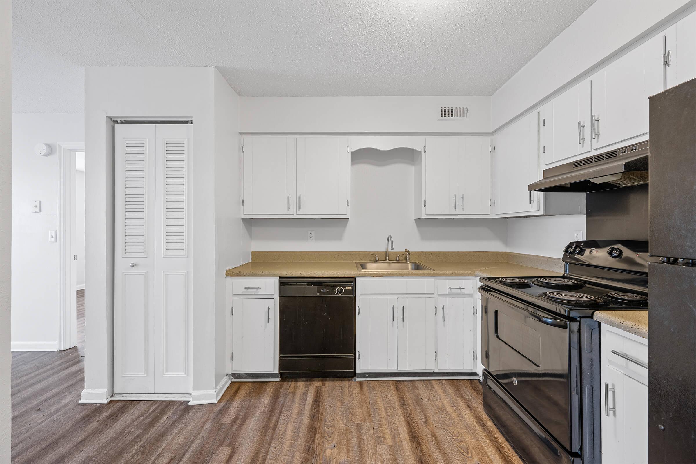 A clean, modern kitchen featuring white cabinets, a stainless steel sink, a black dishwasher, and a black stove and oven. The countertop is light-colored, and the floor has dark wood laminate. A closet is visible on the left, and the space has a bright and spacious feel.