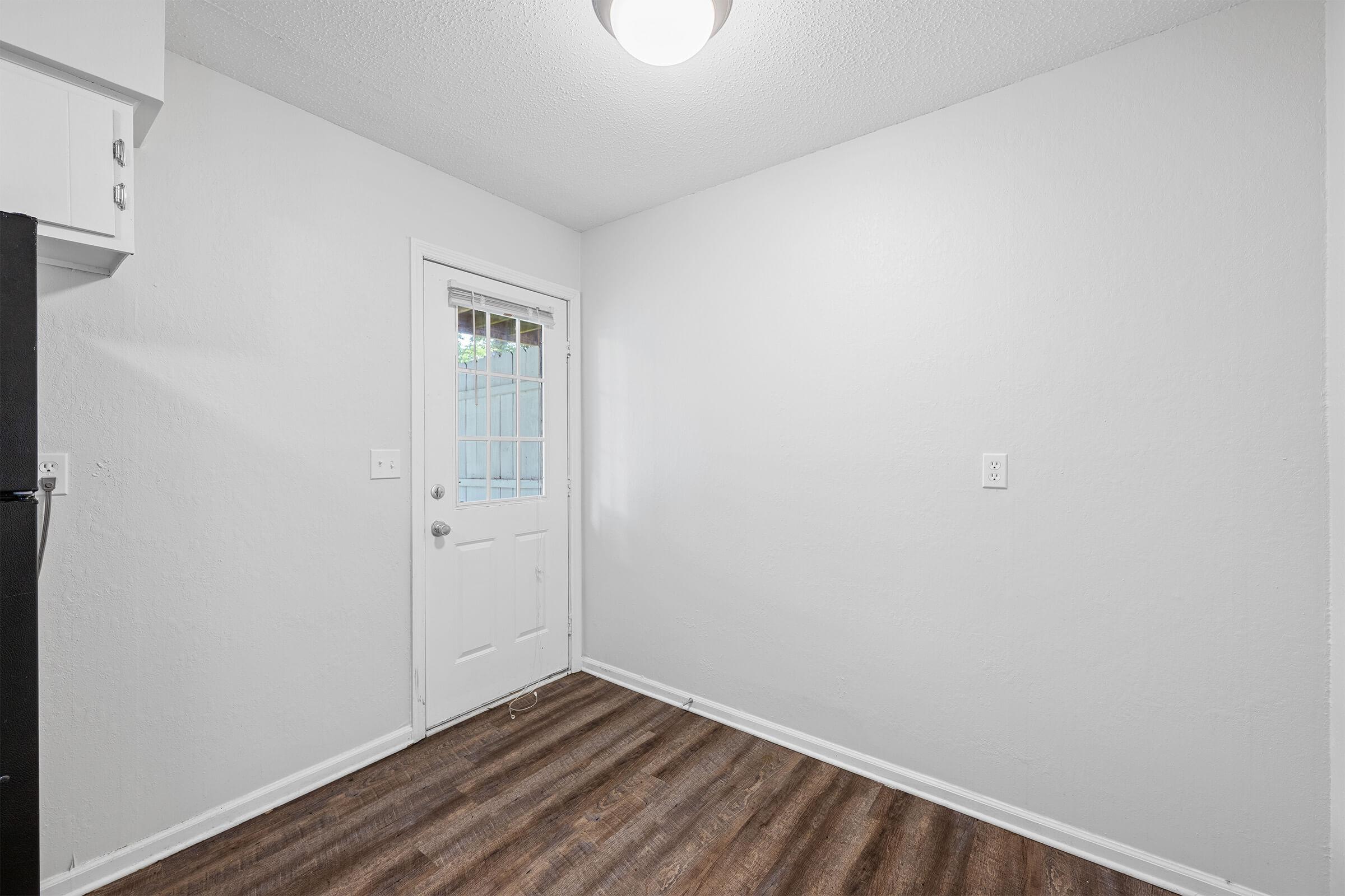 A small, empty room with light gray walls and dark wooden flooring. There is a white door leading outside on the right side, and a window nearby. A ceiling light is visible, illuminating the space, which has no furniture or decor. A black refrigerator is partially seen on the left.