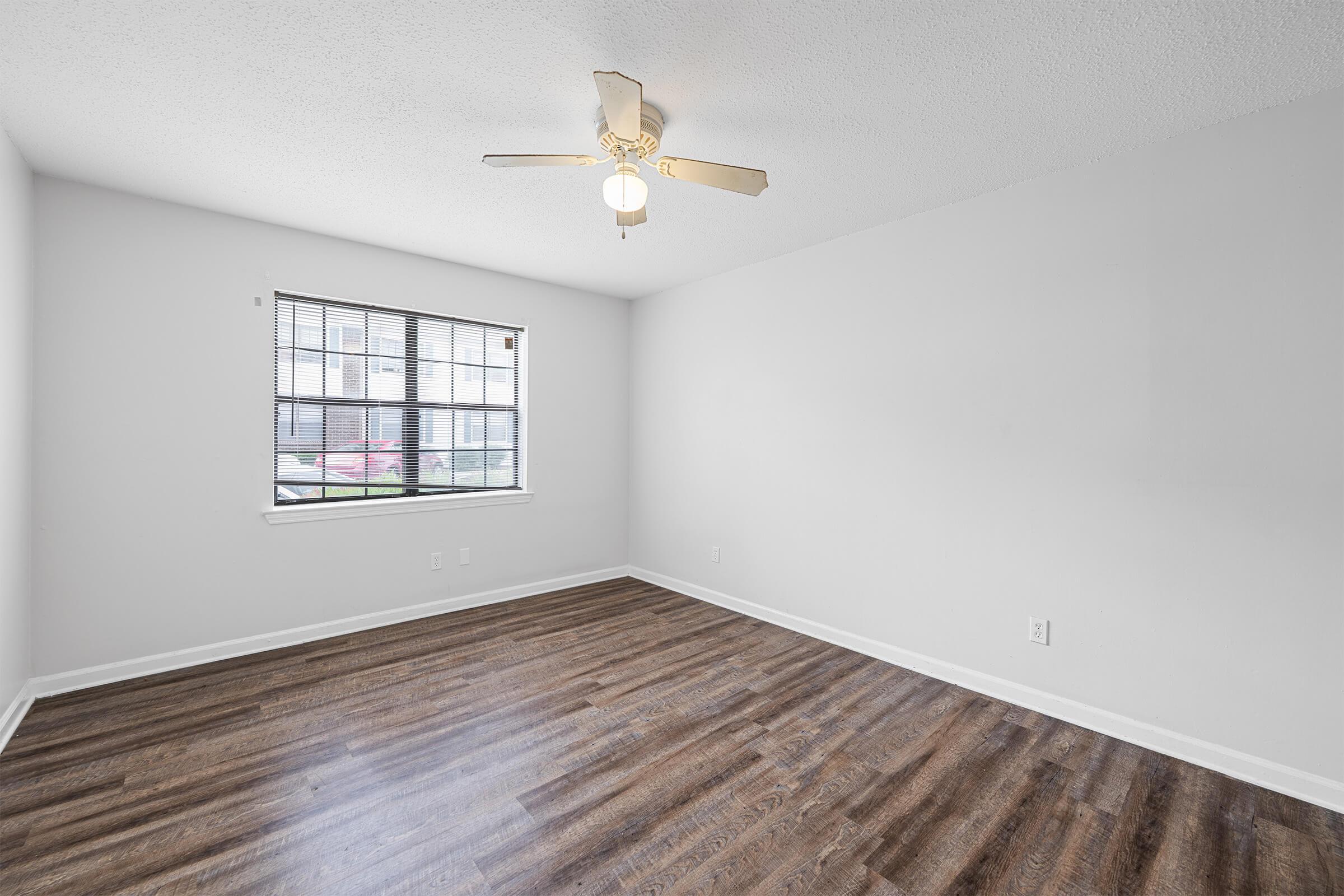 A vacant room with light gray walls, a ceiling fan with light fixture, and brown laminate flooring. There is a window with blinds allowing natural light to enter. The space is clean and empty, ready for furniture or decoration.