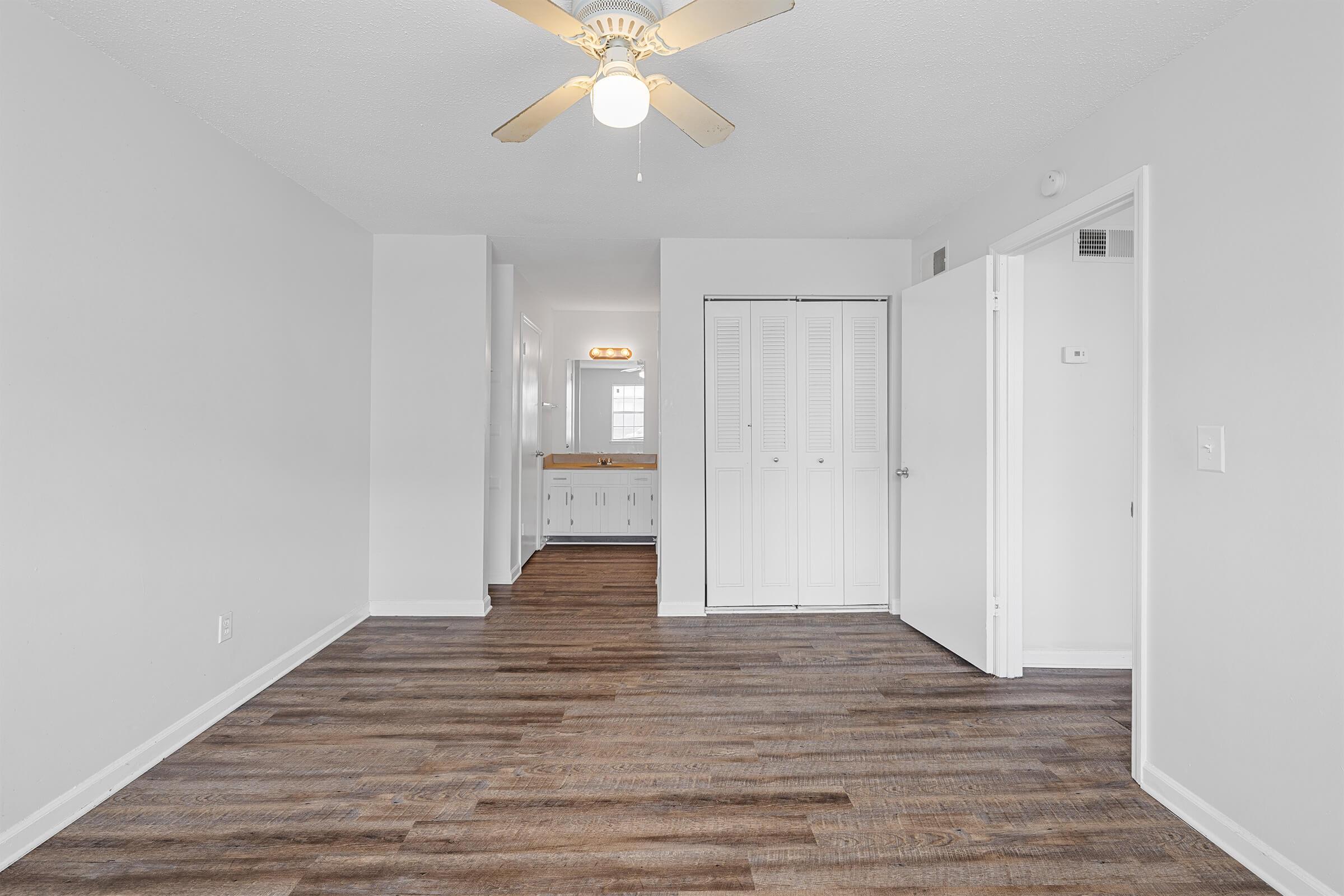 A bright, open living space with light-colored walls and wooden flooring. The room features a ceiling fan and a closet with sliding doors. To the left, there is an entryway leading to another room with a countertop and additional storage. The space is well-lit and inviting.