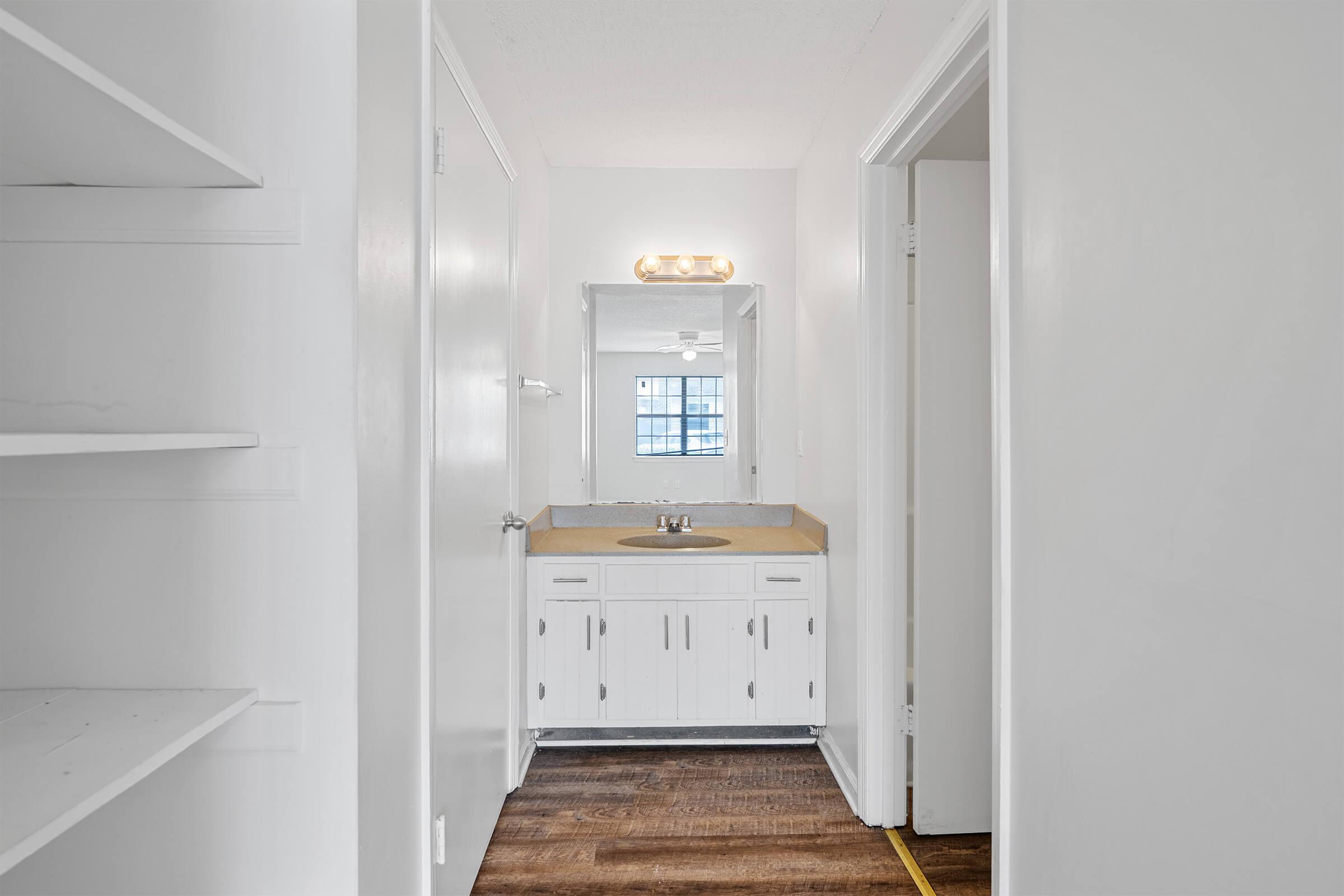 A well-lit bathroom area featuring a vanity with a sink, white cabinetry, a large mirror, and light fixtures. The space has light-colored walls and flooring, with a shelf on the left and doors leading to other areas, creating an open and clean atmosphere.