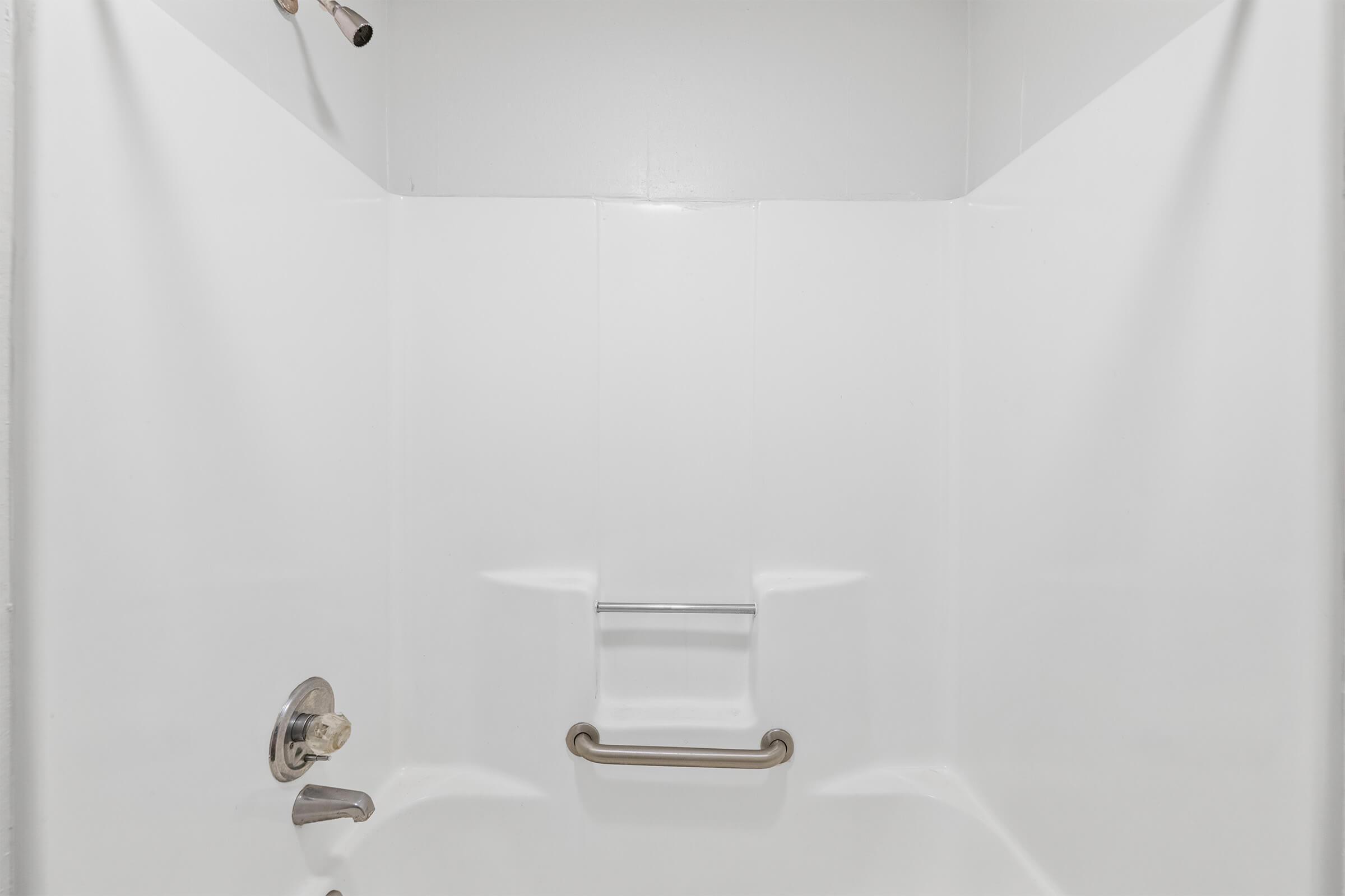 A clean, empty shower stall featuring white walls, a metal grab bar, and a simple faucet. The space is well-lit and minimalistic, highlighting the functional design with no decorative elements present.