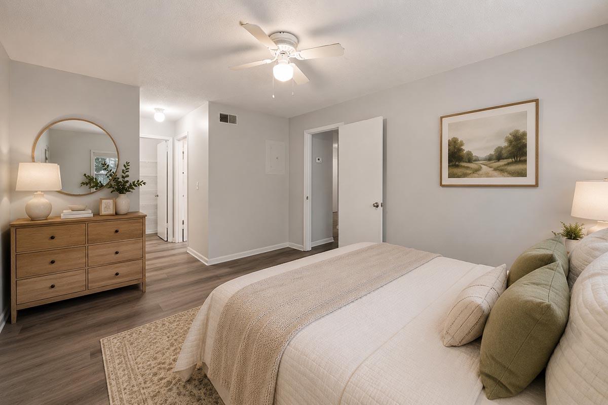 A cozy bedroom featuring a queen-sized bed with neutral bedding, a soft green throw pillow, and a decorative wall painting. A round mirror is mounted above a wooden dresser, which holds a lamp and decor. The room has light gray walls, wooden flooring, and a ceiling fan, contributing to a warm, inviting atmosphere.