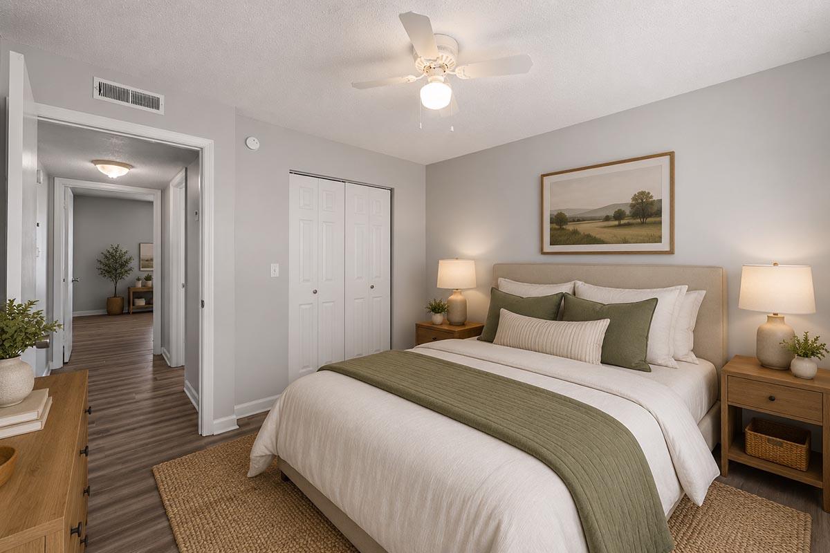 A cozy bedroom featuring a queen-sized bed with a light-colored duvet and green accent pillows. Two bedside tables with lamps flank the bed. A landscape painting adorns the wall above the bed, and natural light fills the space. The room has a neutral color palette and wood flooring, creating a warm atmosphere.