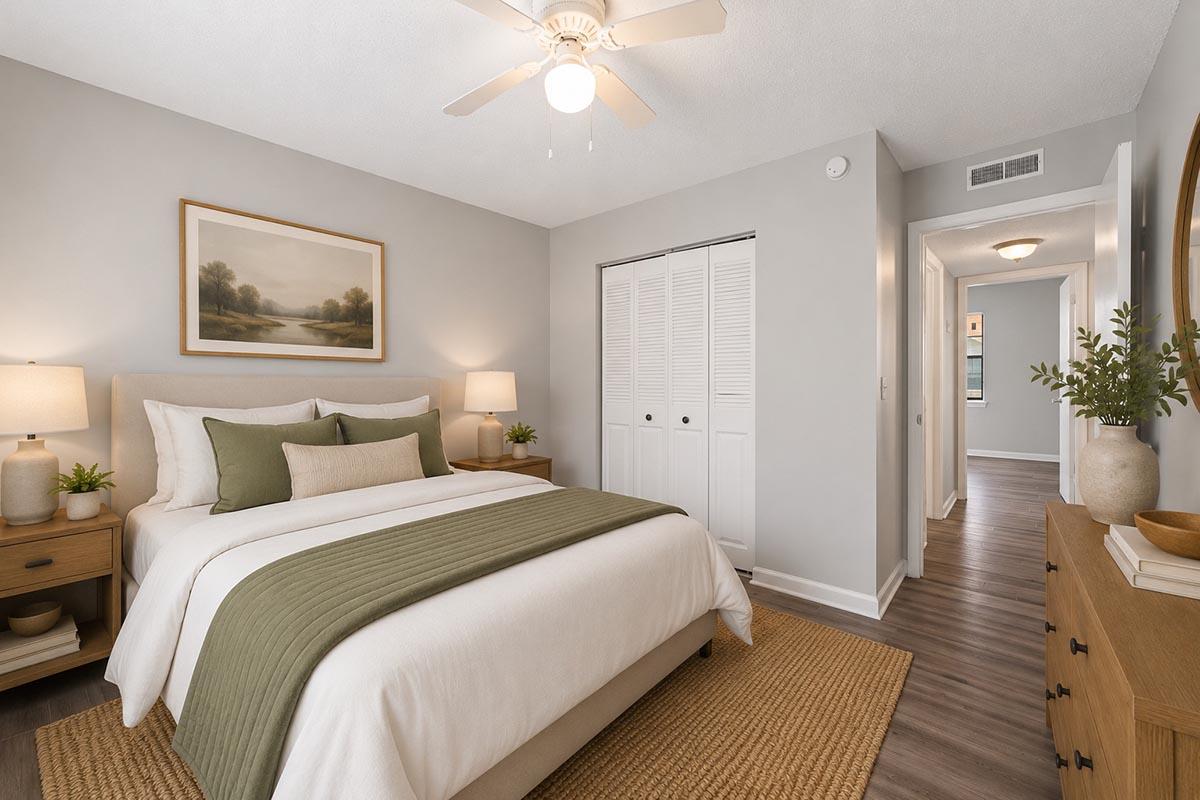 Cozy bedroom featuring a queen-sized bed with green and white bedding, two bedside lamps, and a wooden nightstand. A large painting of a landscape hangs above the bed. The room has light gray walls, a ceiling fan, and a closet with double doors, with a natural fiber rug on the hardwood floor.